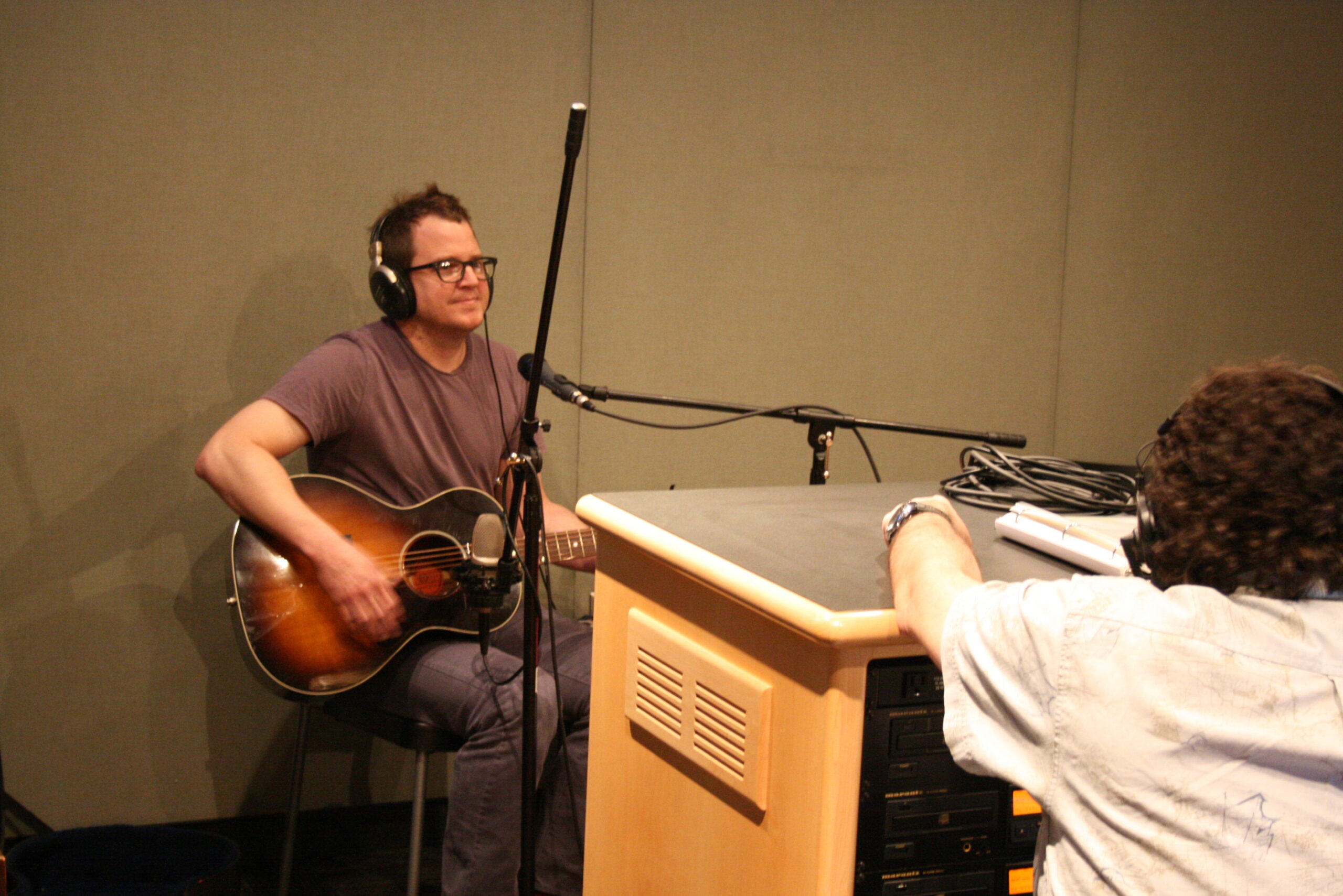 An adult wearing headphones holding an acoustic guitar being interviewed by an adult wearing headphones in a radio studio