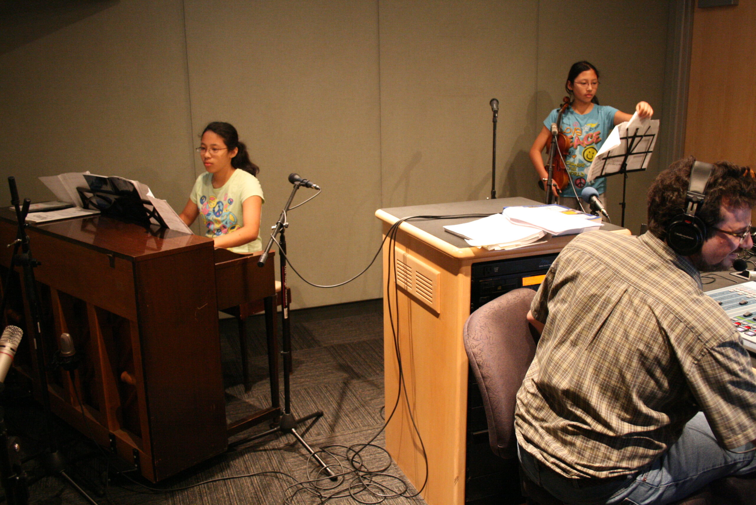 A teen playing a piano and a teen holding a violin as an adult wearing headphones listens in a radio studio