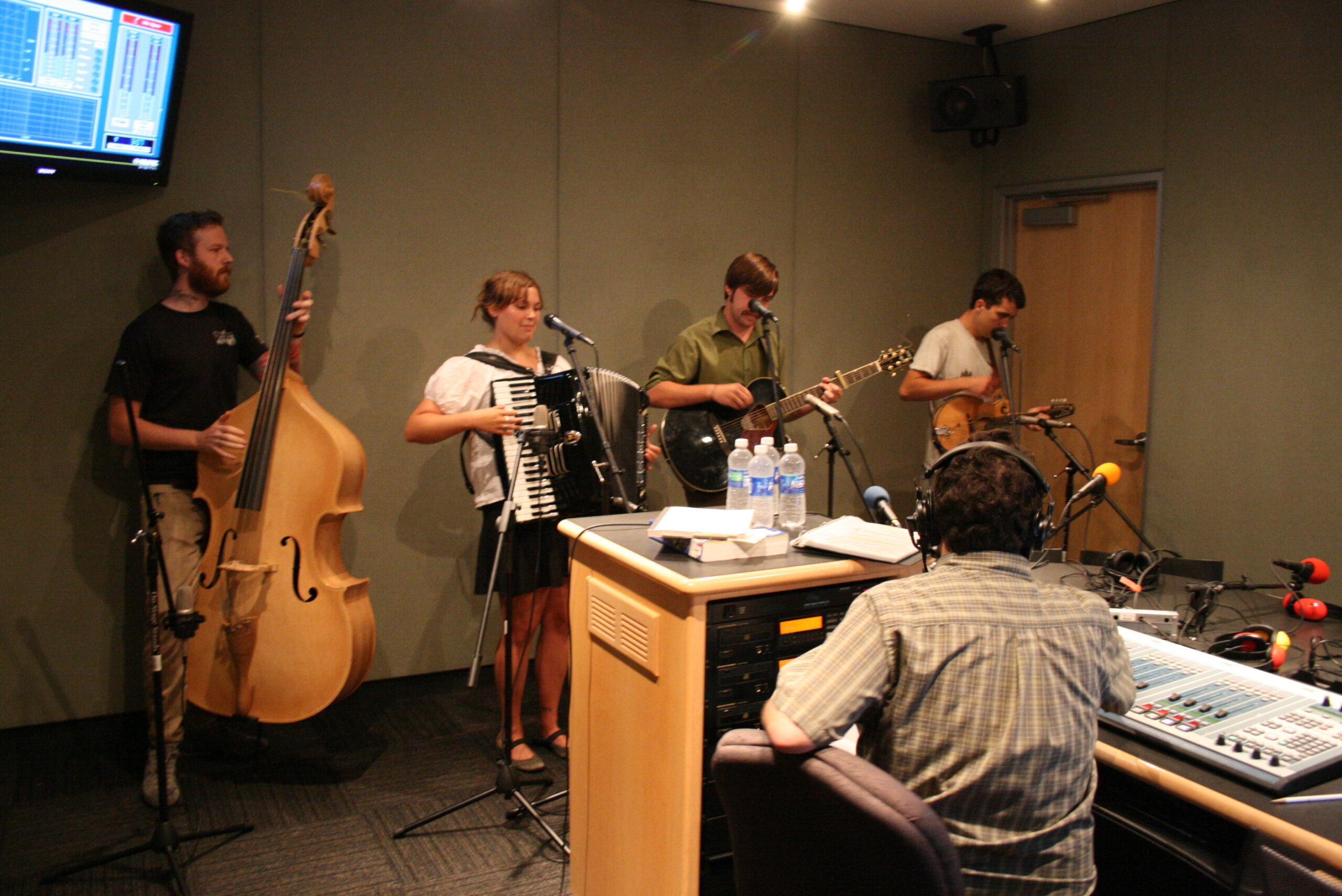 An adult playing an upright bass, an adult playing an accordion, an adult playing an acoustic guitar, and an adult playing a mandolin as an adult wearing headphones listens in a radio studio