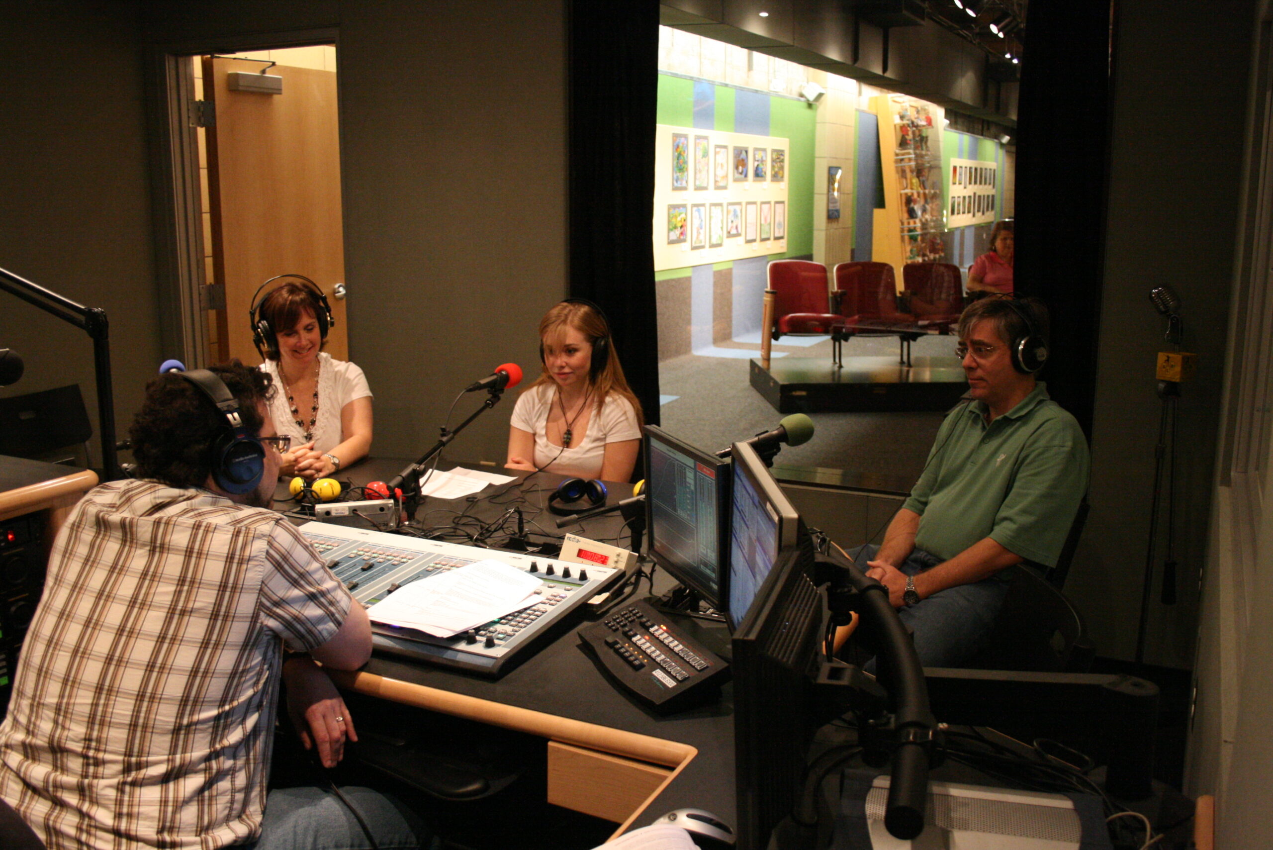 Two adults and a teen wearing headphones sitting behind microphones being interviewed by an adult wearing headphones sitting behind an audio mixer in a radio studio