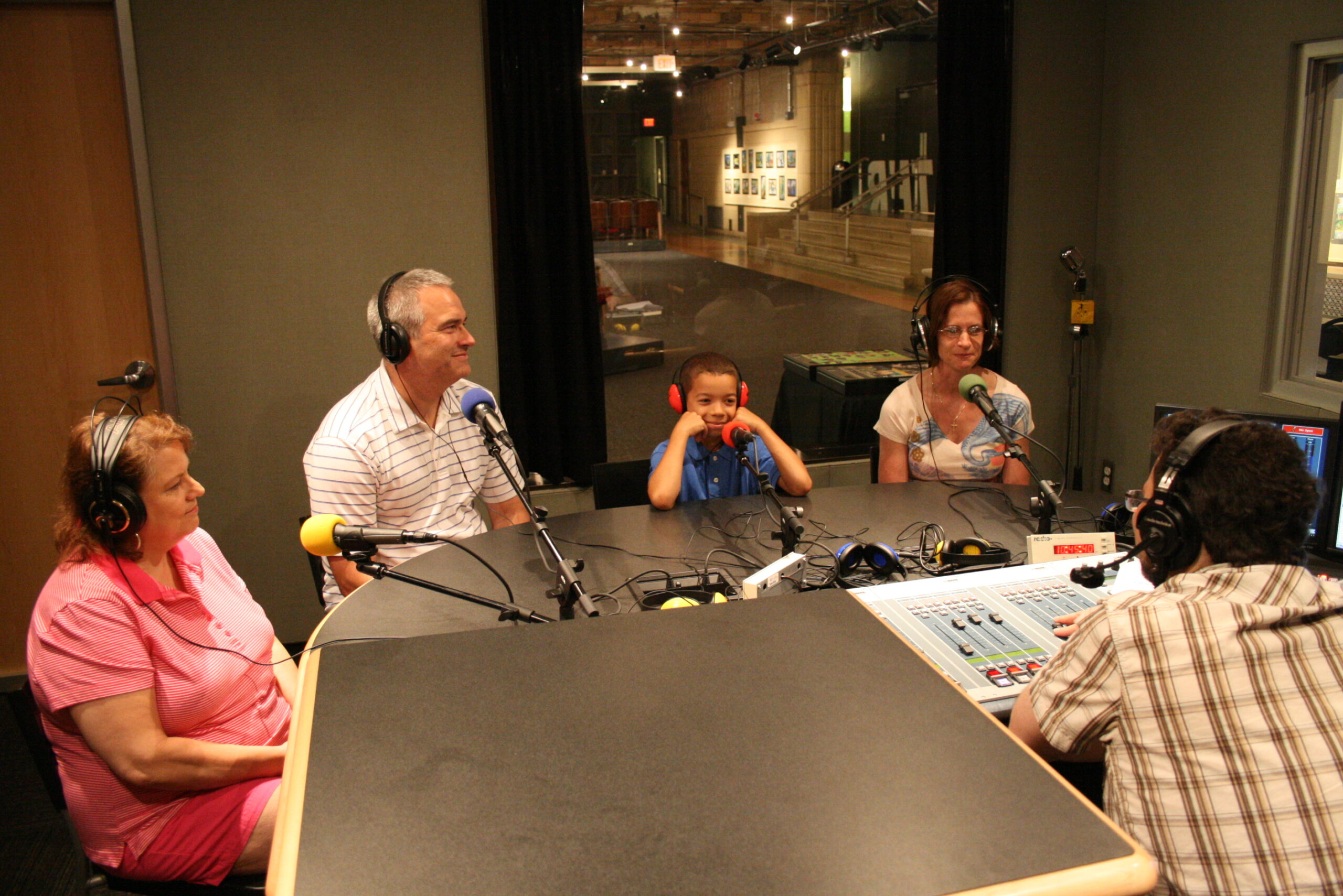 Three adults and a youth wearing headphones speaking into microphones being interviewed by an adult wearing headphones sitting behind an audio mixer in a radio studio