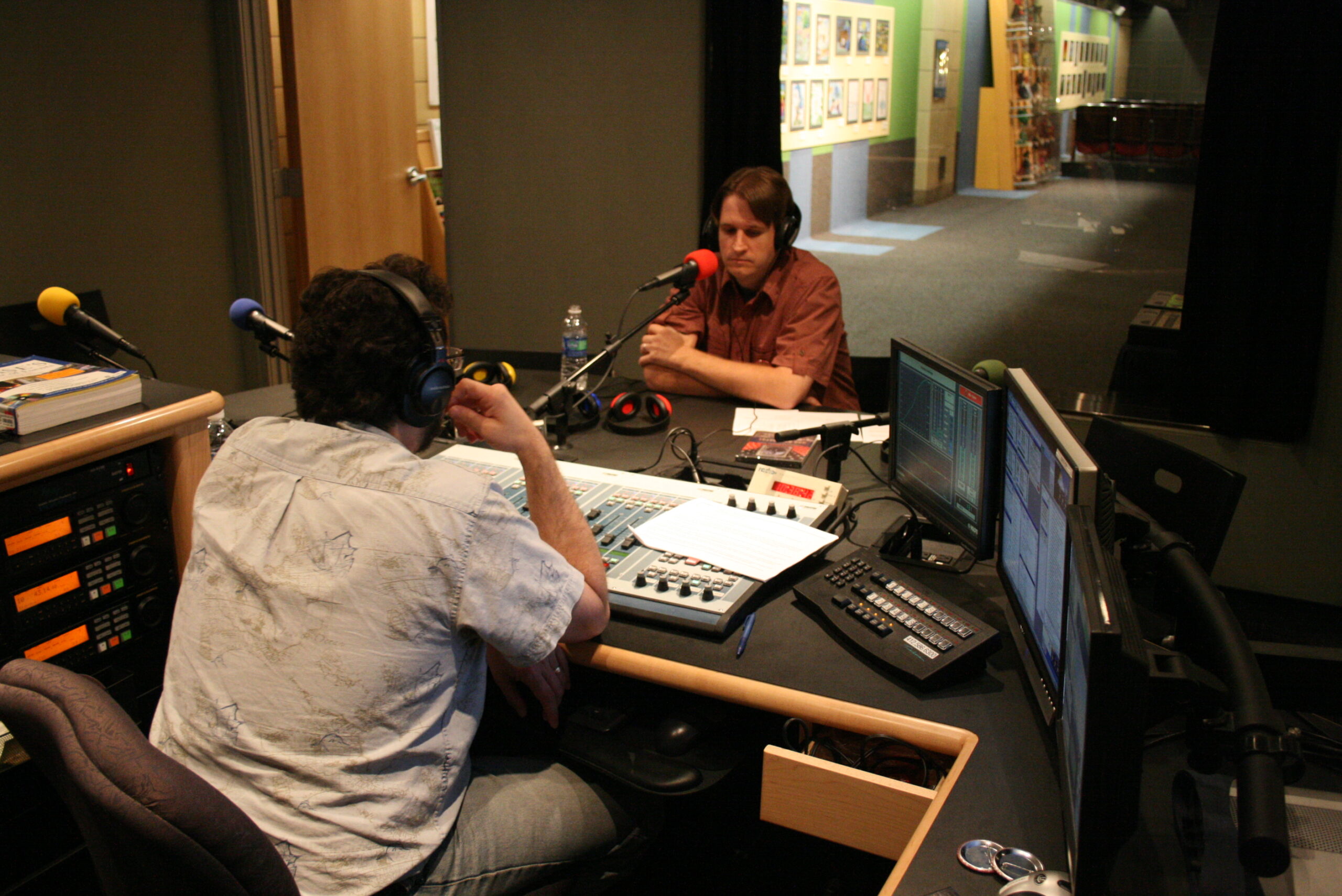 An adult wearing headphones sitting behind a microphone being interviewed by an adult wearing headphones sitting behind an audio mixer in a radio studio