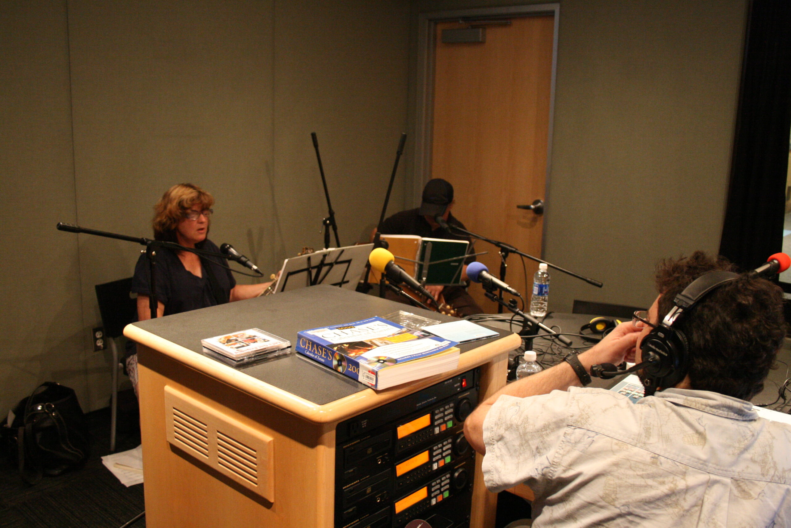 Two adults sitting and holding stringed instruments being interviewed by an adult wearing headphones in a radio studio