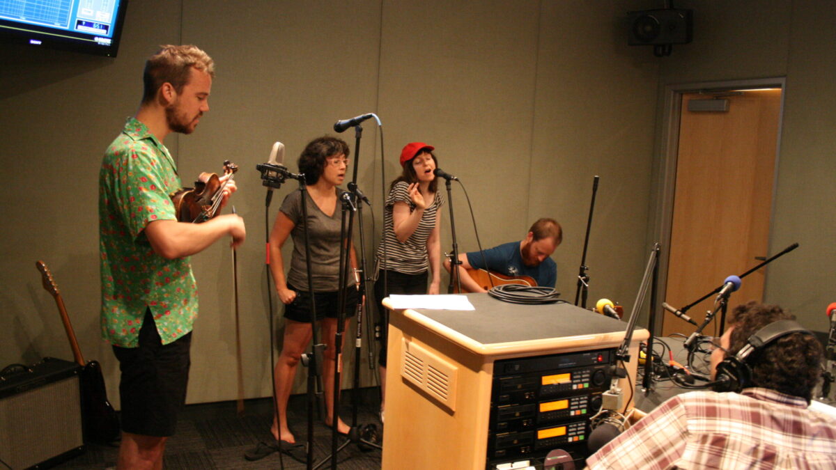 An adult playing a ukulele, two adults singing into microphones, and an adult playing an acoustic guitar in a radio studio