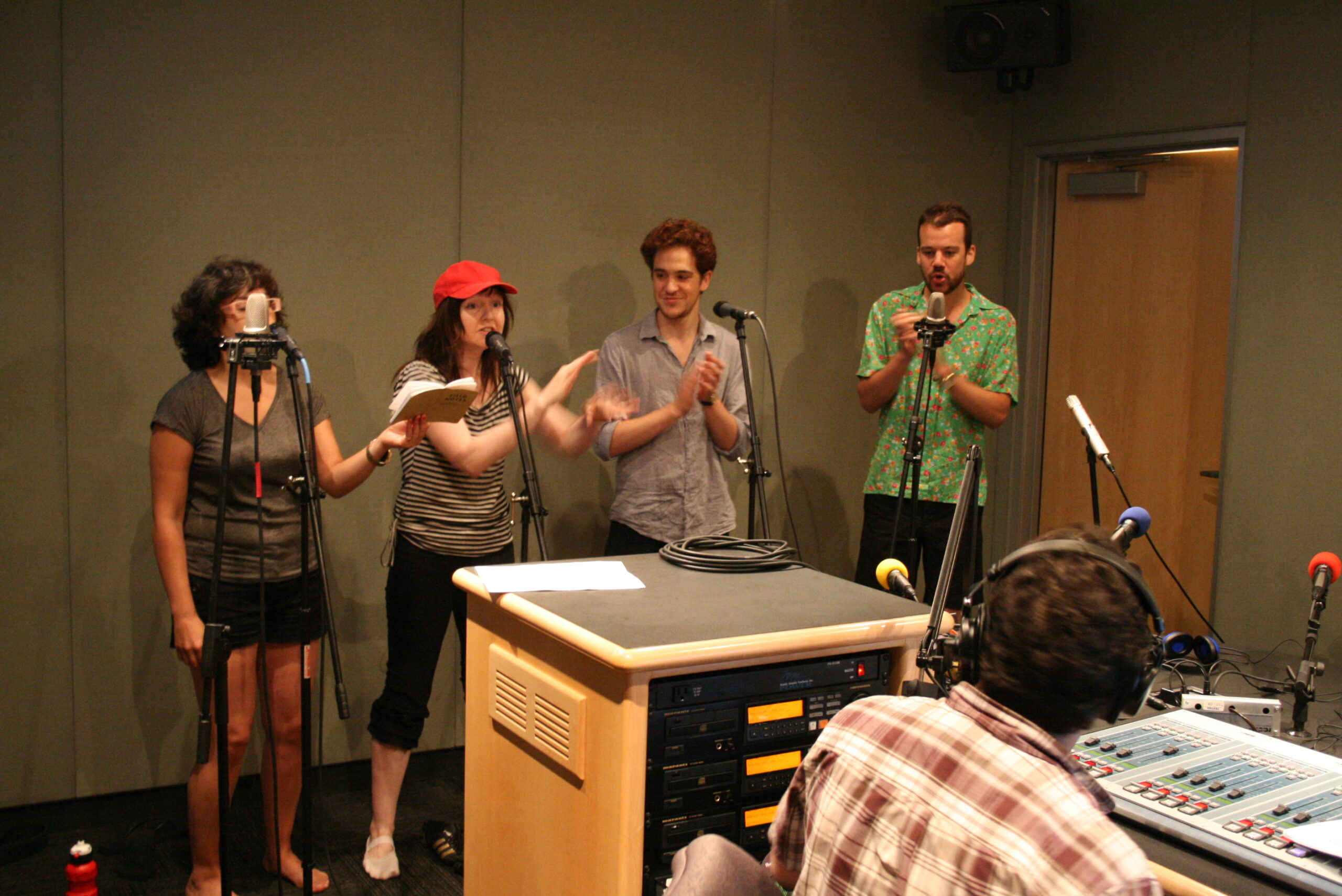 Four adults singing in front of microphones in a radio studio