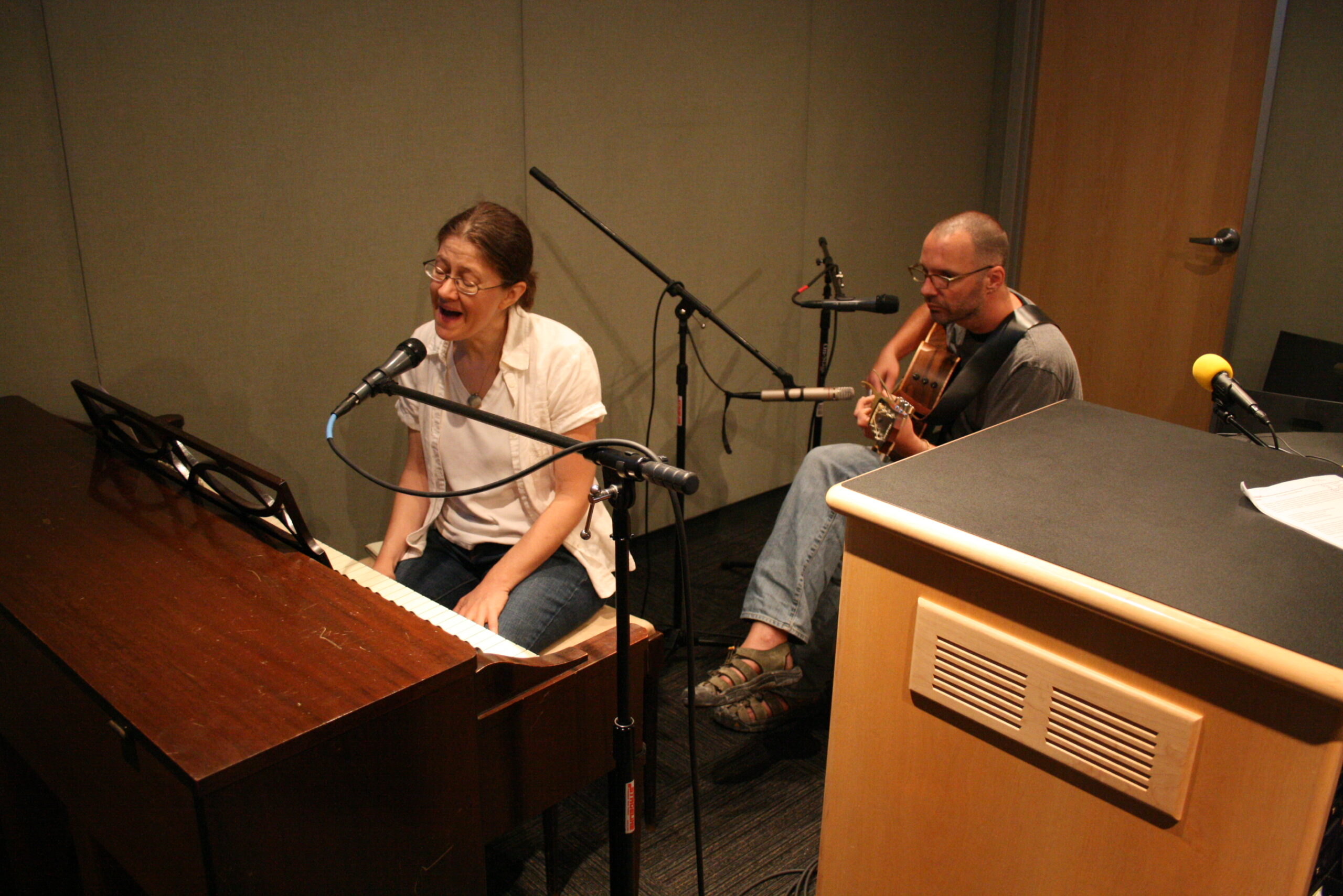 An adult singing into a microphone and playing a piano and an adult playing an acoustic guitar in a radio studio