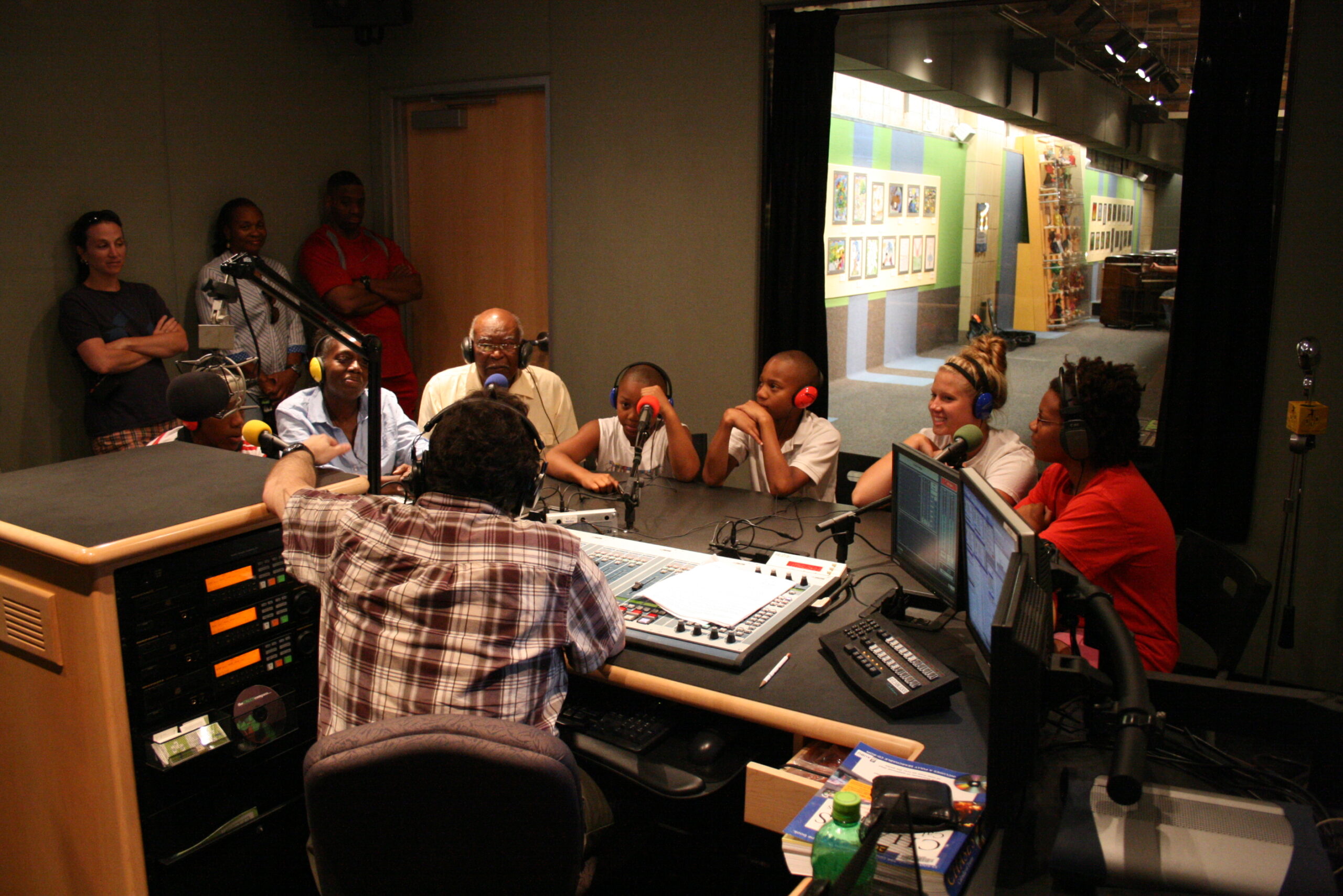 Four adults and three youth wearing headphones speaking into microphones being interviewed by an adult wearing headphones sitting behind an audio mixer as three adults stand and watch in a radio studio