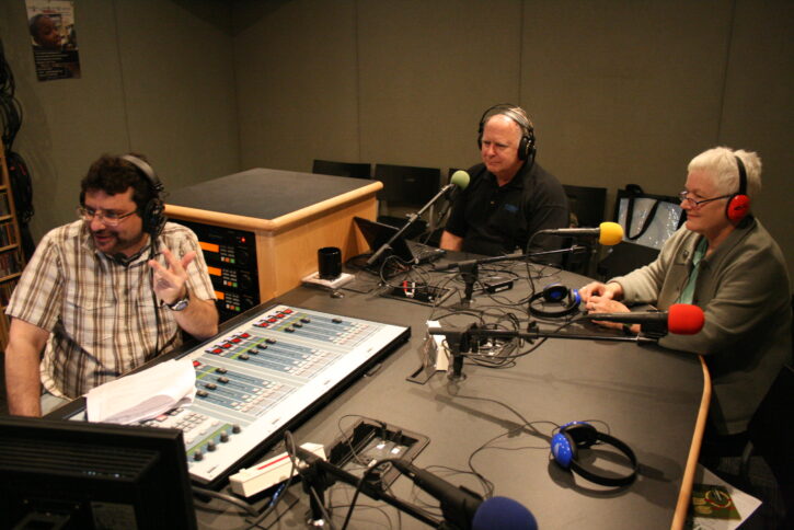 Two adults wearing headphones speaking into microphones being interviewed by an adult wearing headphones sitting behind an audio mixer in a radio studio