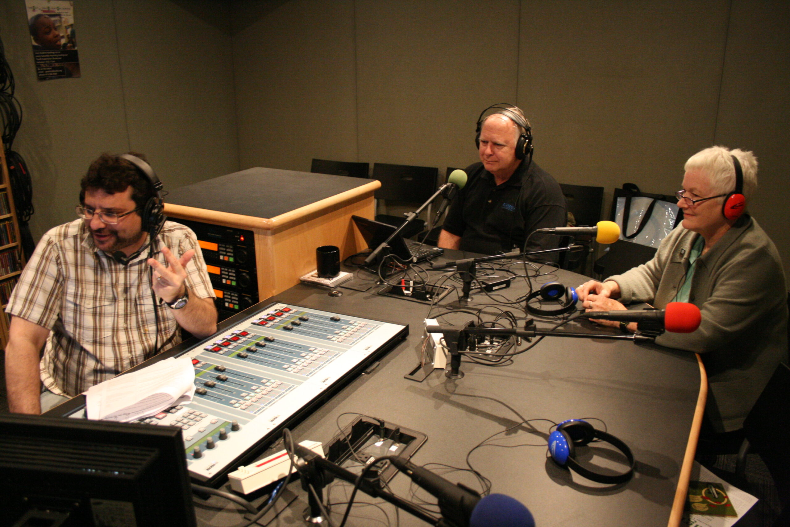 Two adults wearing headphones speaking into microphones being interviewed by an adult wearing headphones sitting behind an audio mixer in a radio studio