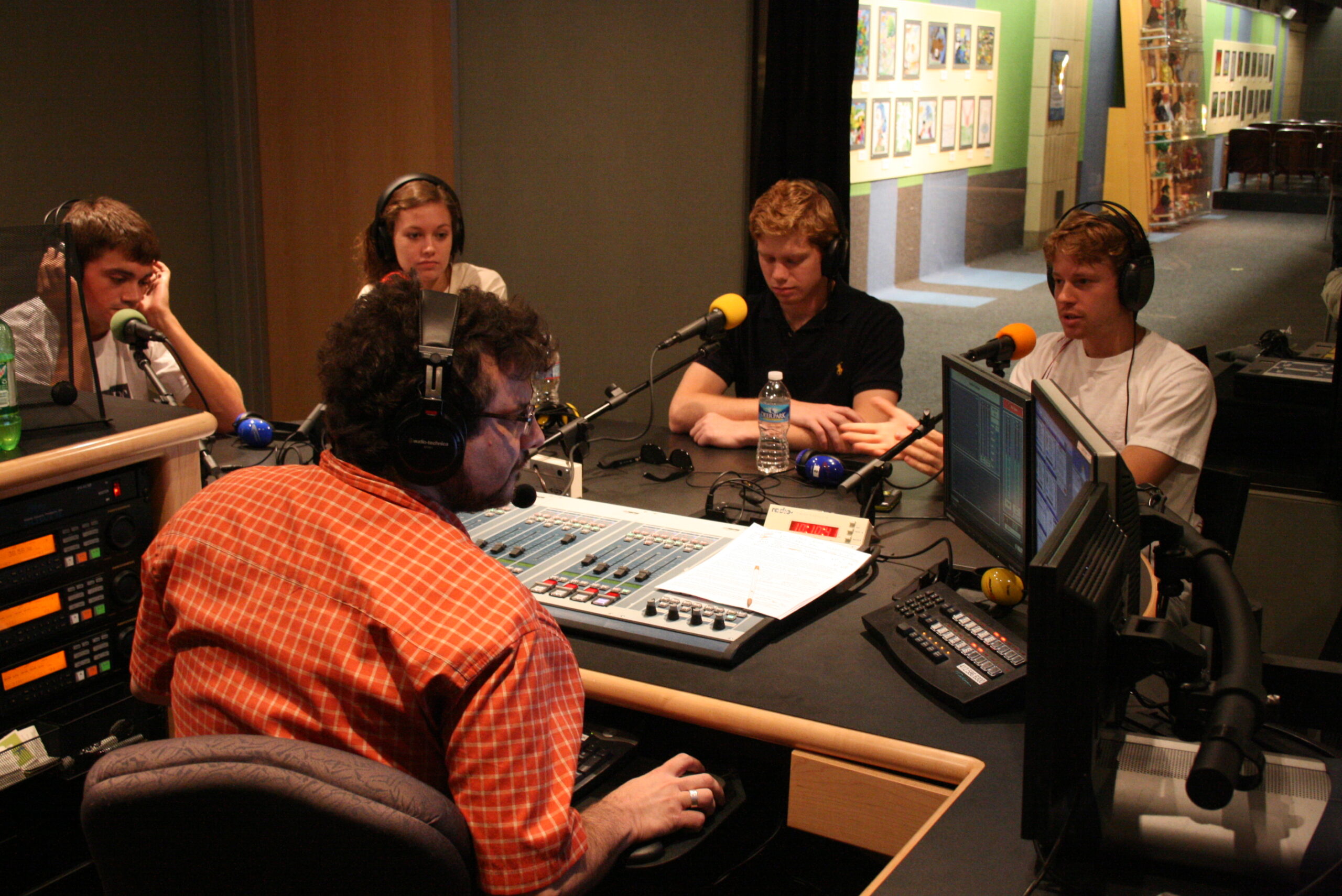Four teens wearing headphones sitting behind microphones being interviewed by an adult wearing headphones sitting behind an audio mixer in a radio studio
