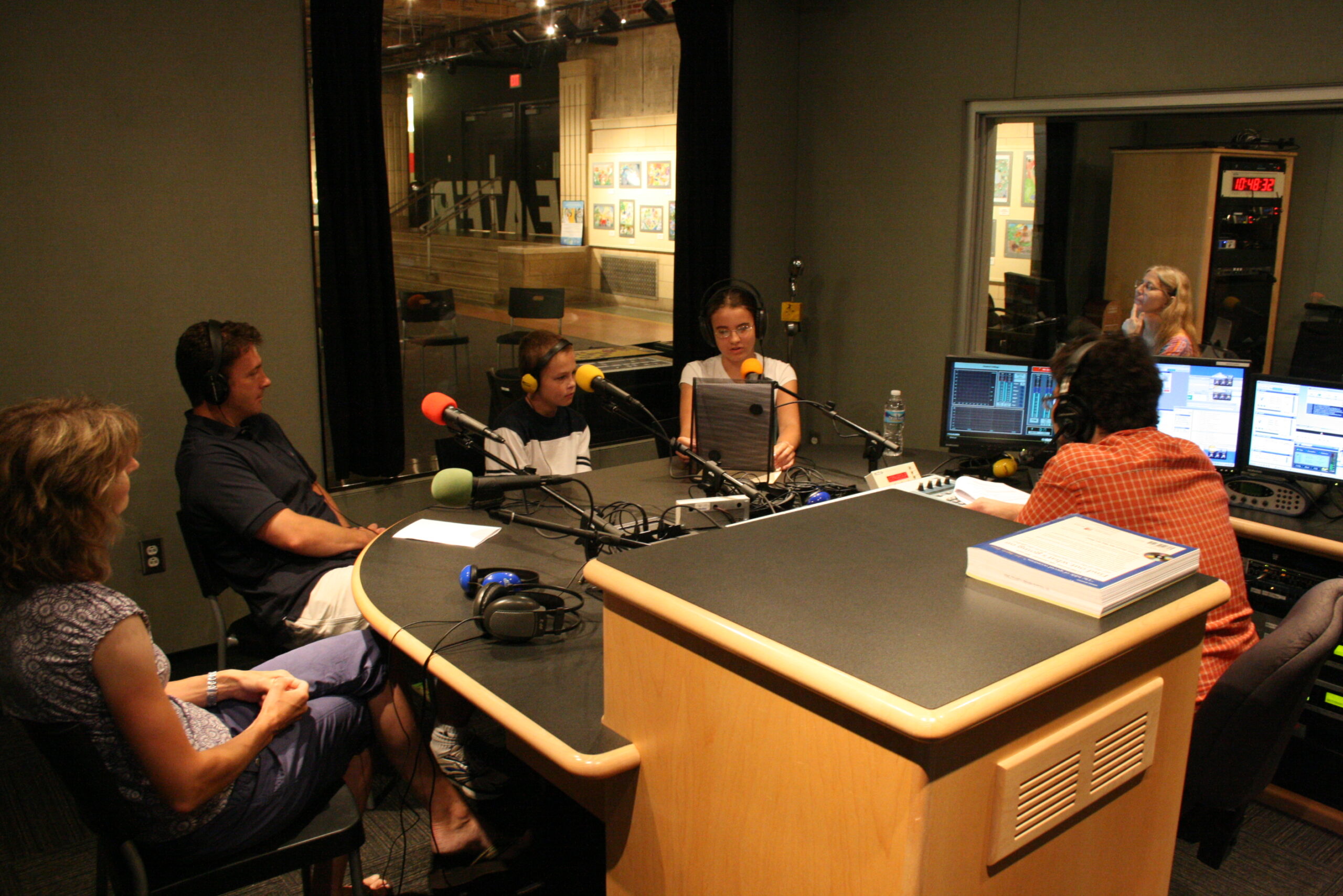 Two youth wearing headphones and two adults speaking into microphones being interviewed by an adult wearing headphones in a radio studio