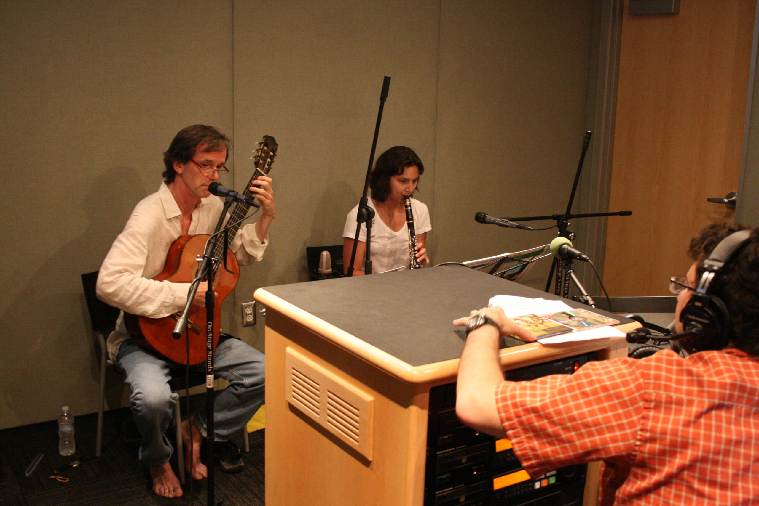 An adult playing an acoustic guitar and an adult playing a clarinet as an adult wearing headphones listens in a radio studio