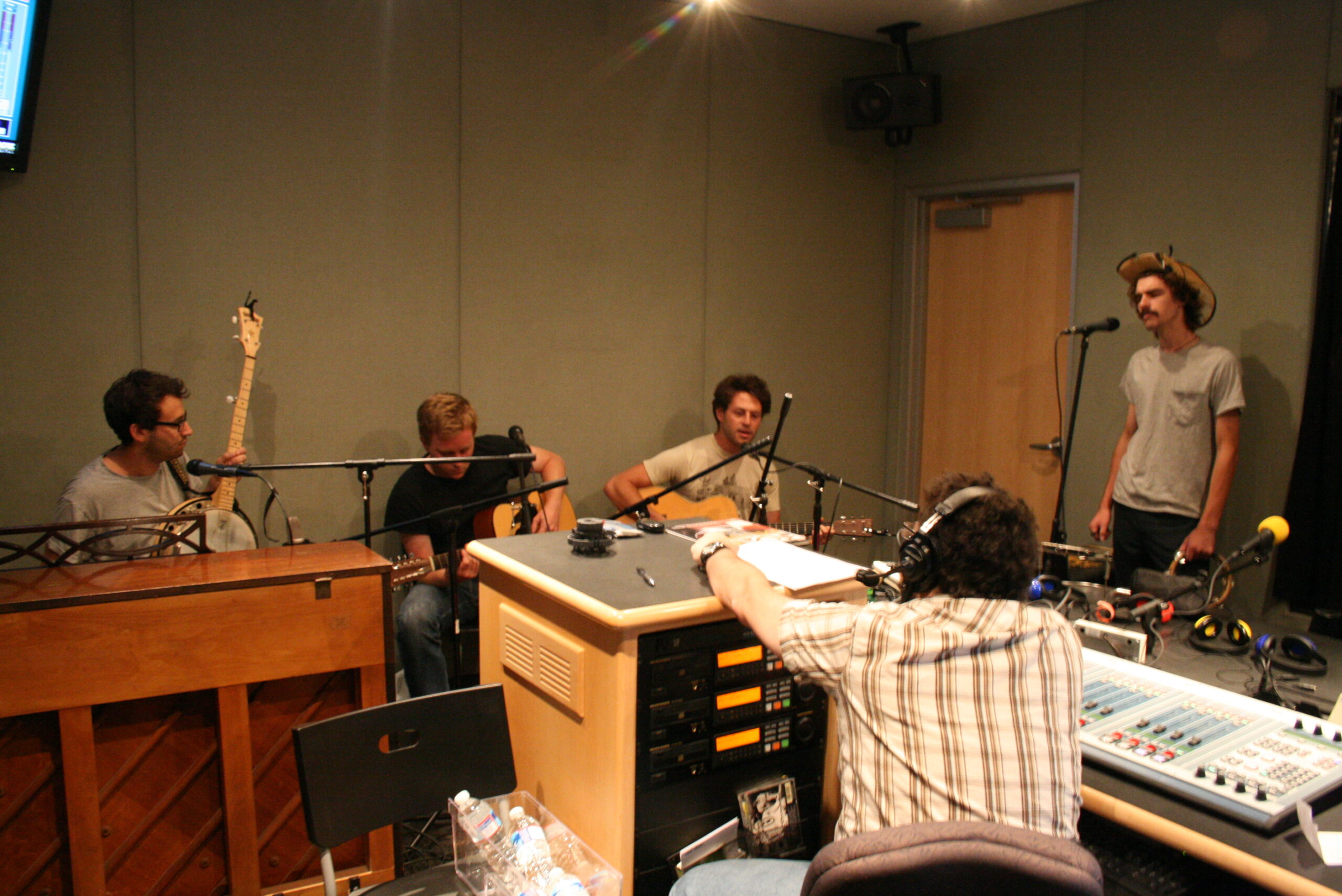 An adult playing an acoustic guitar, two adults playing acoustic guitars, and an adult singing into a microphone as an adult wearing headphones listens in a radio studio