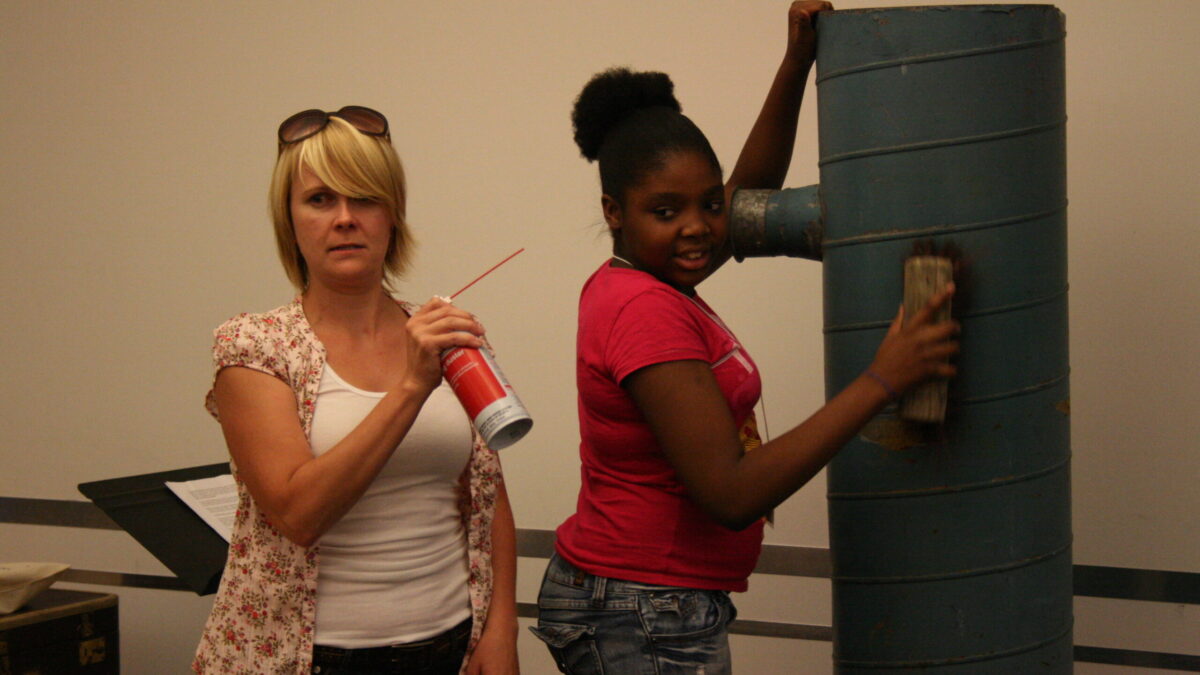 SDA 005 An adult holding a spray can and a youth holding a brush standing next to a gray cylinder