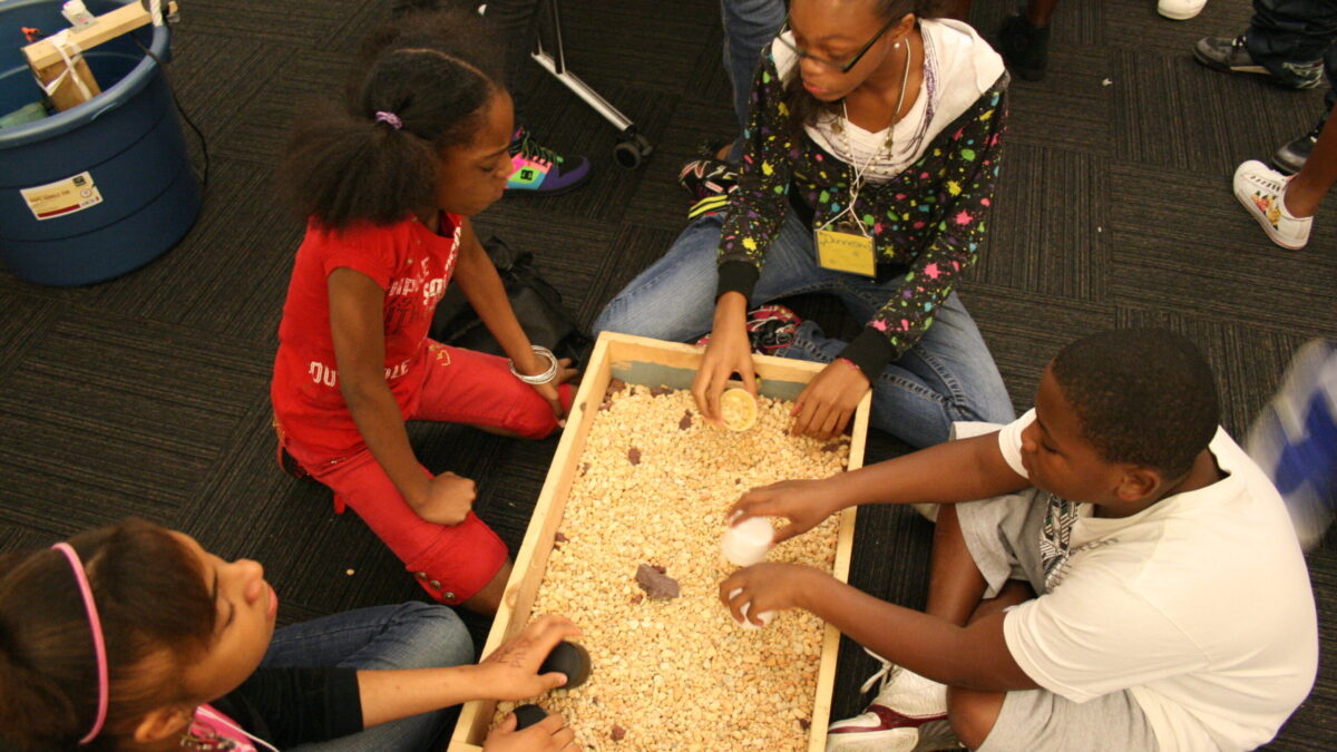 SDA 015 Four youth sitting around a wooden box filled with pebbles in a classroom