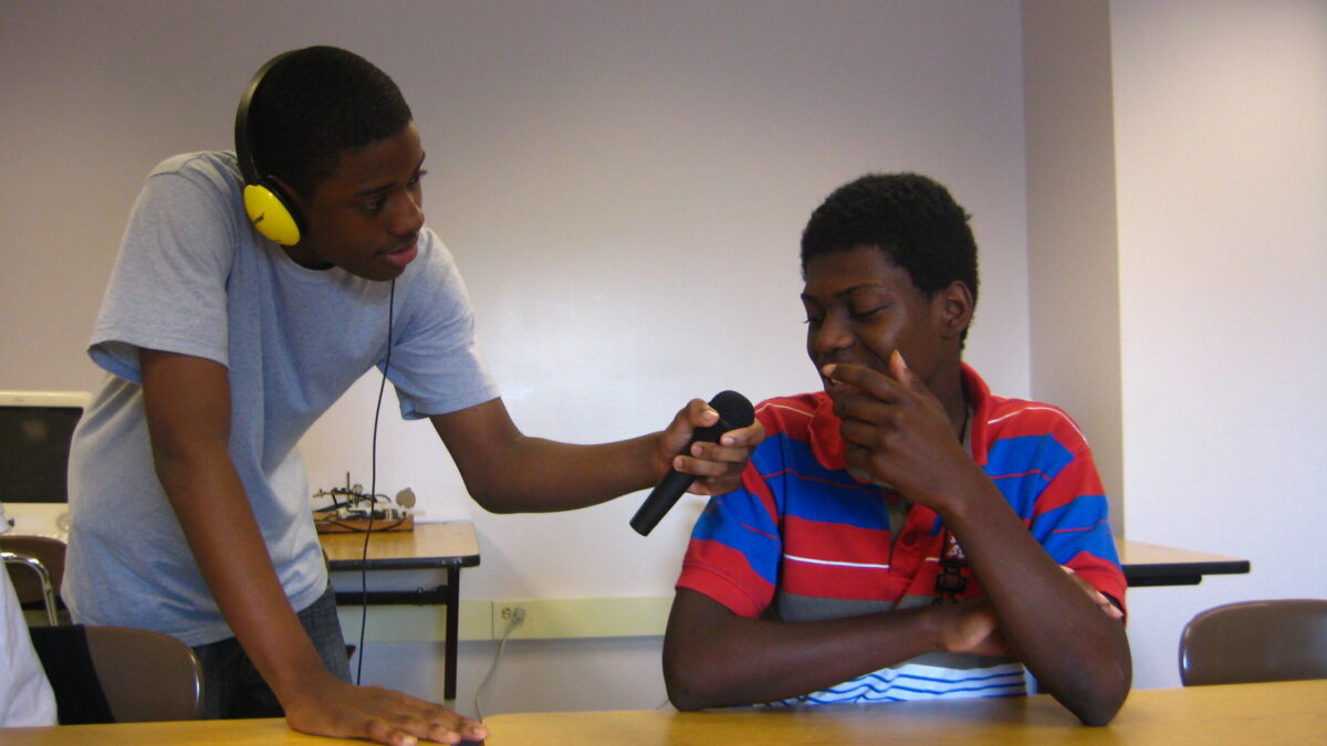 A teen wearing headphones holding a microphone for a teen in a classroom