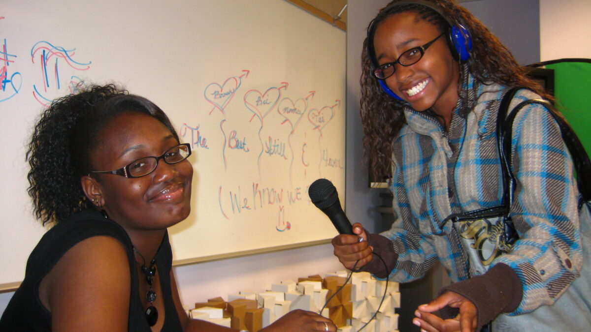 A teen wearing headphones smiling and holding a microphone for an adult in a classroom