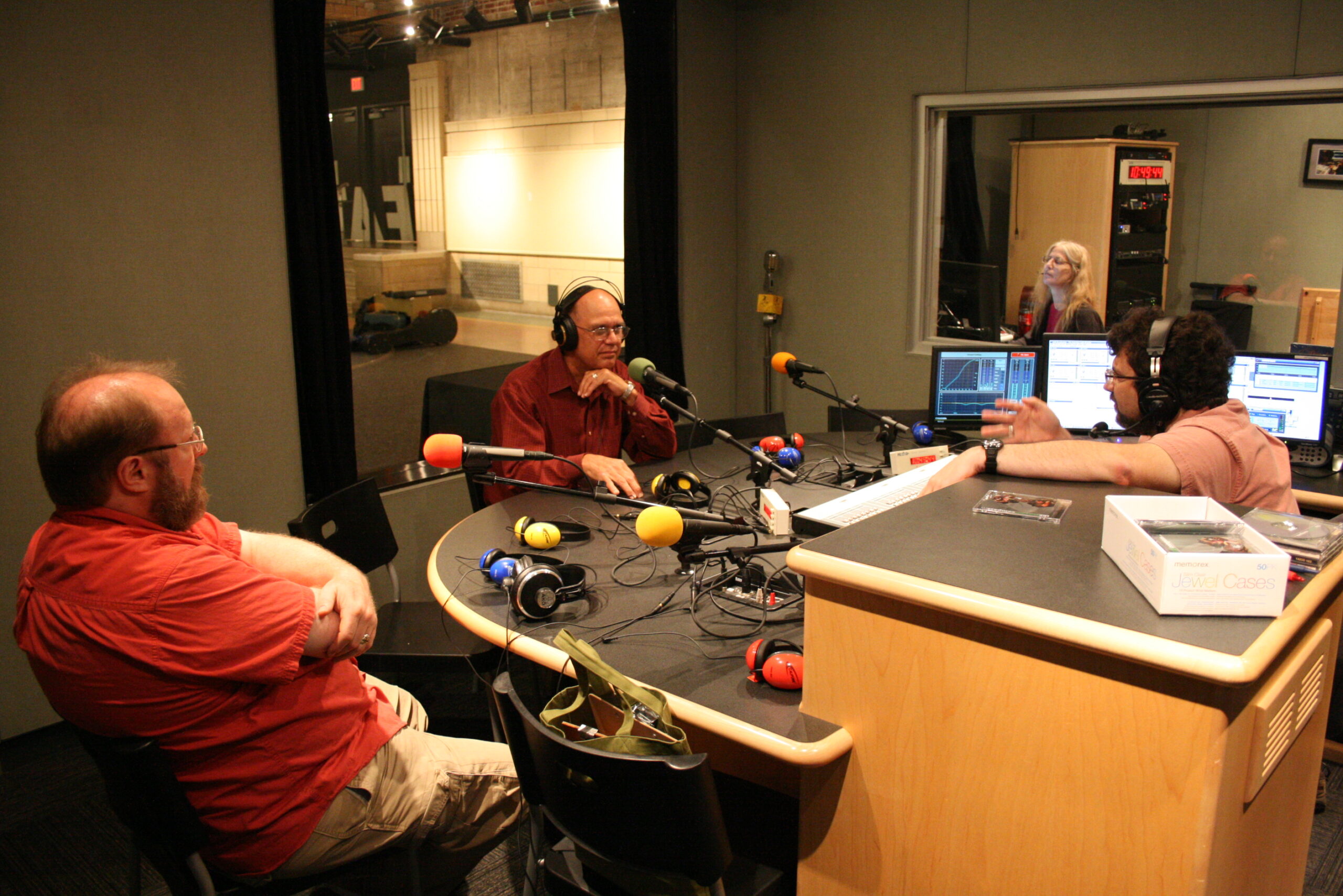 Two adults, one wearing headphones, speaking into microphones being interviewed by an adult wearing headphones sitting behind an audio mixer in a radio studio