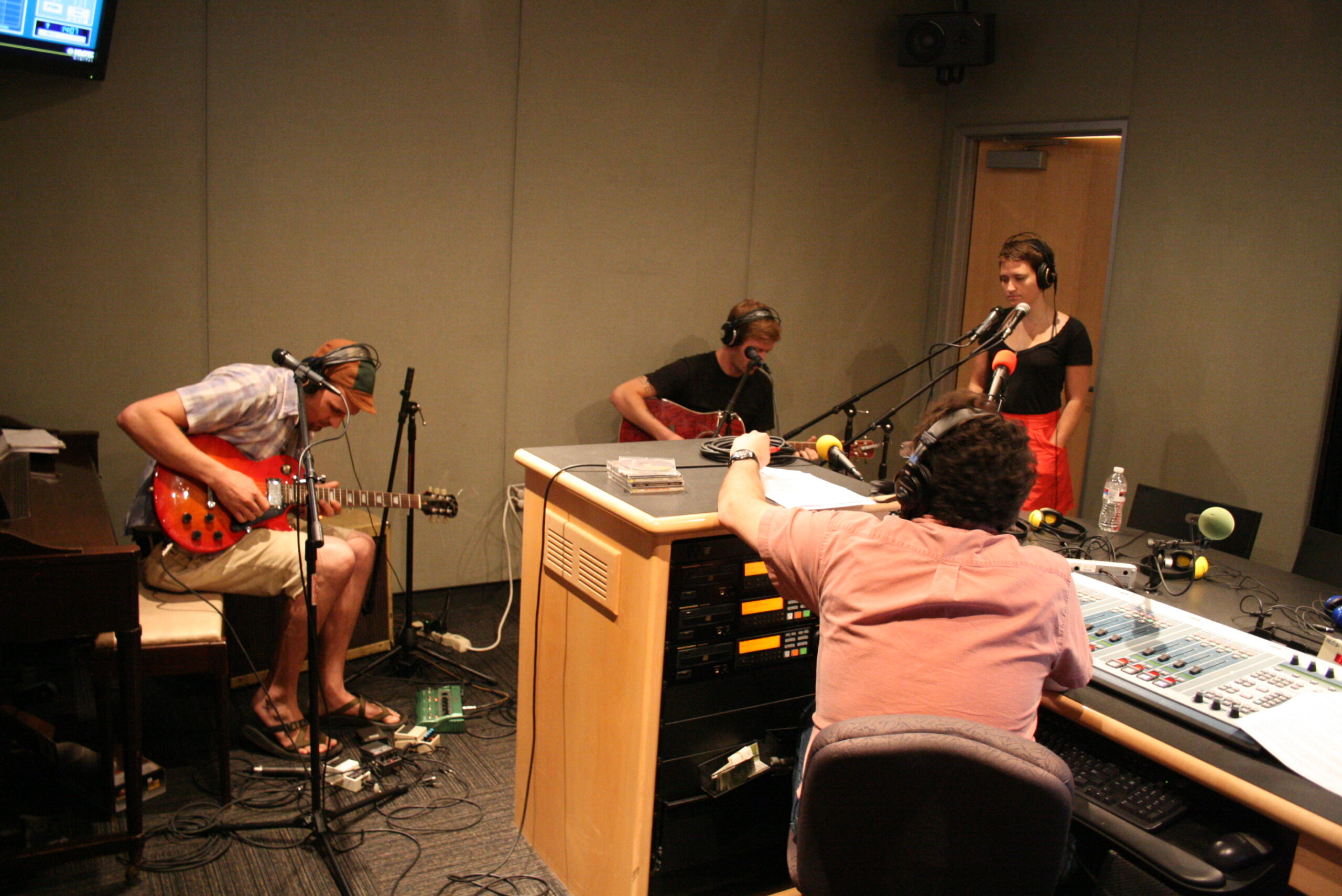 An adult playing an electric guitar, an adult playing an electric bass, and an adult wearing headphones singing into a microphone as an adult wearing headphones listens in a radio studio