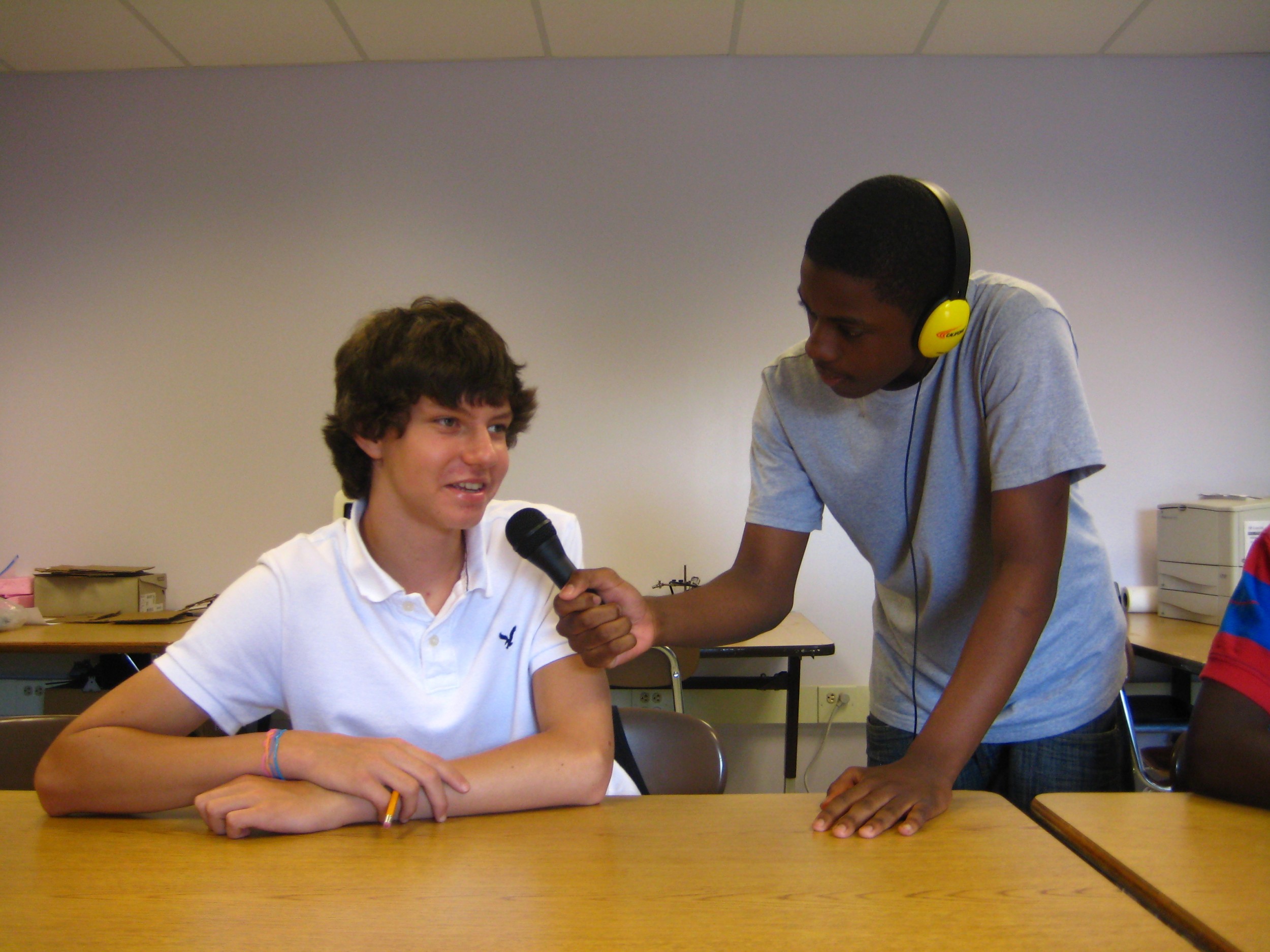 A teen wearing headphones holding a microphone for a teen in a classroom