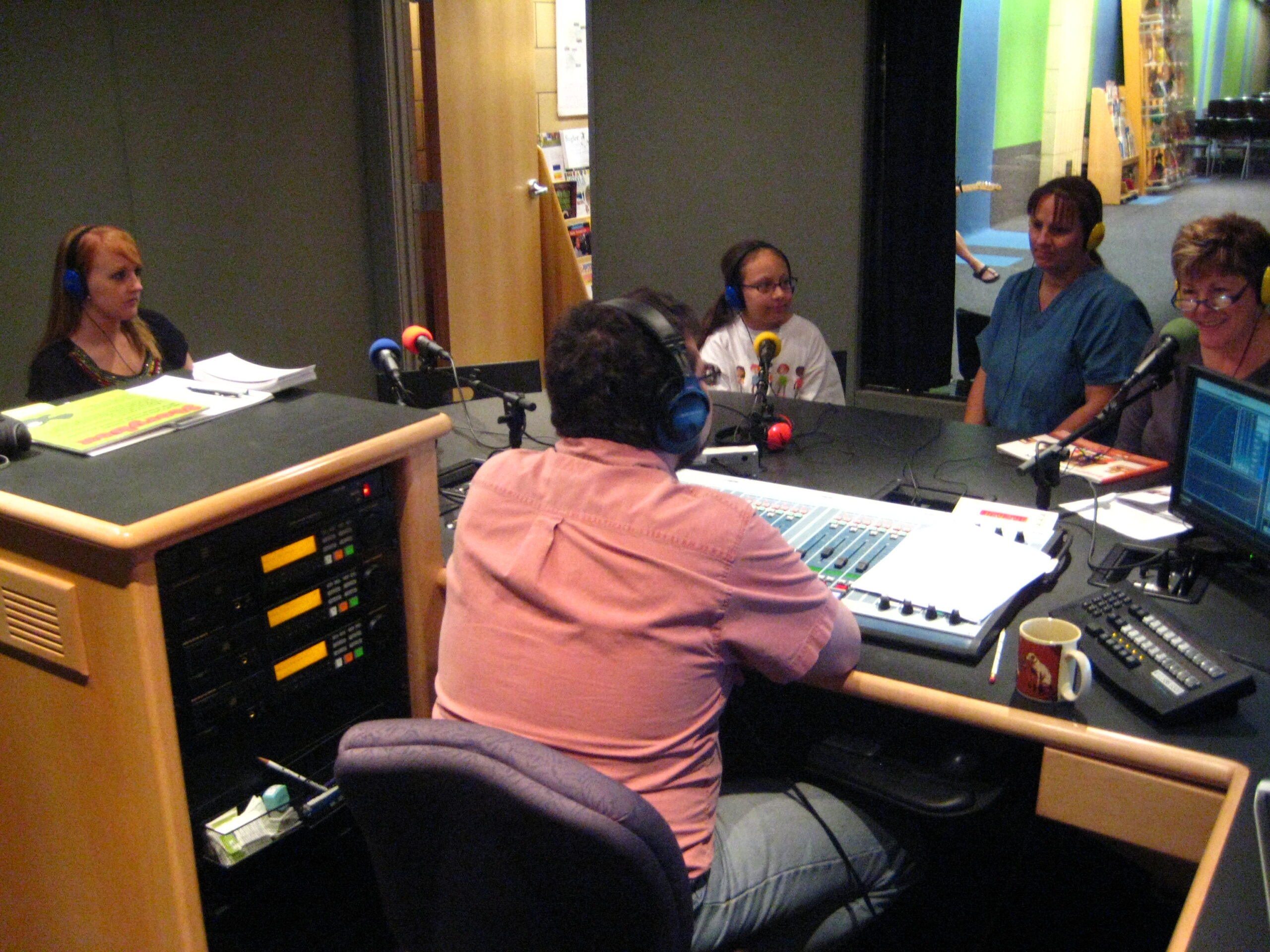 Two adults and a youth wearing headphones speaking into microphones being interviewed by an adult wearing headphones sitting behind an audio mixer in a radio studio