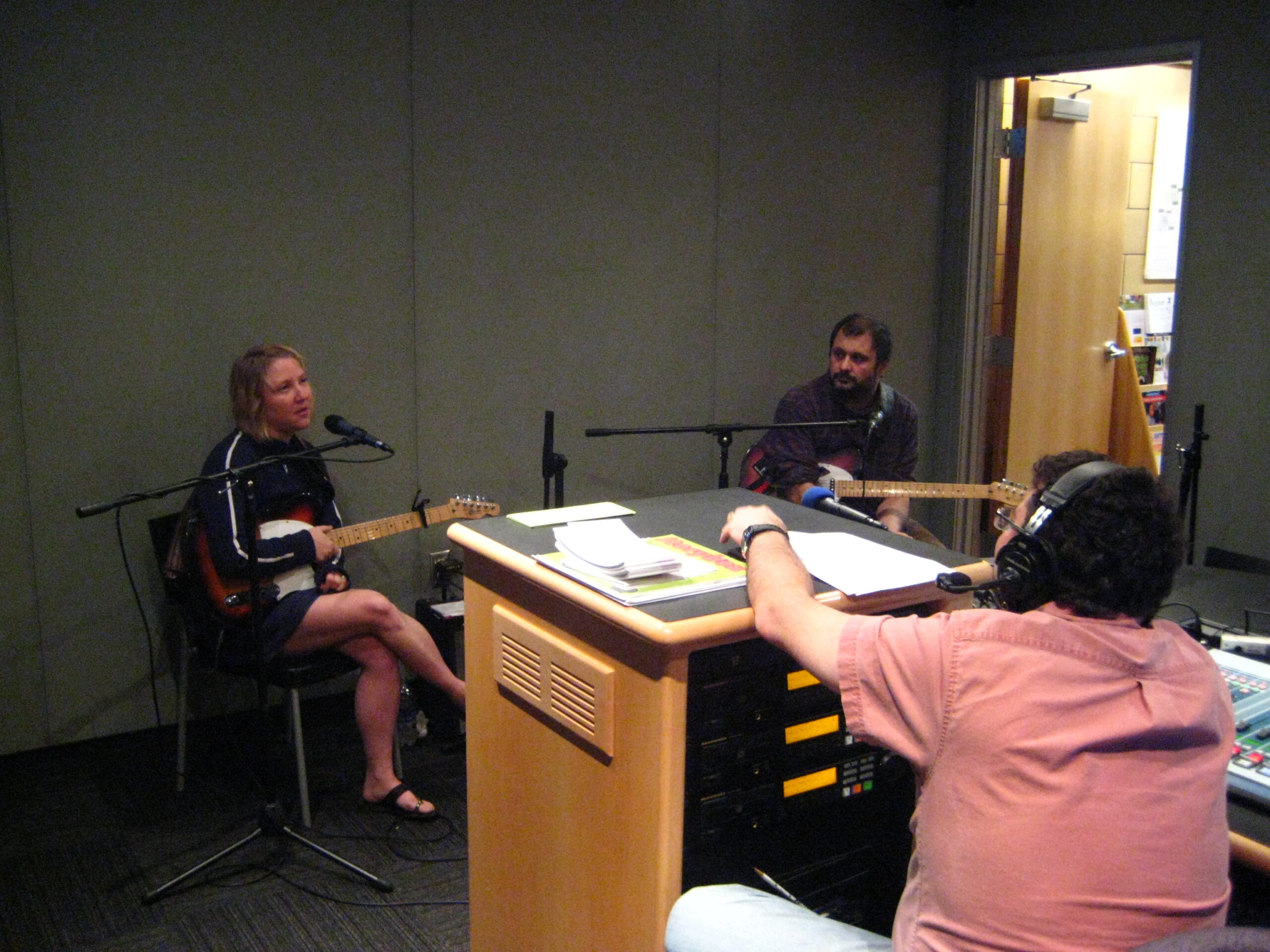 Two adults holding electric guitars and speaking into microphones being interviewed by an adult wearing headphones in a radio studio