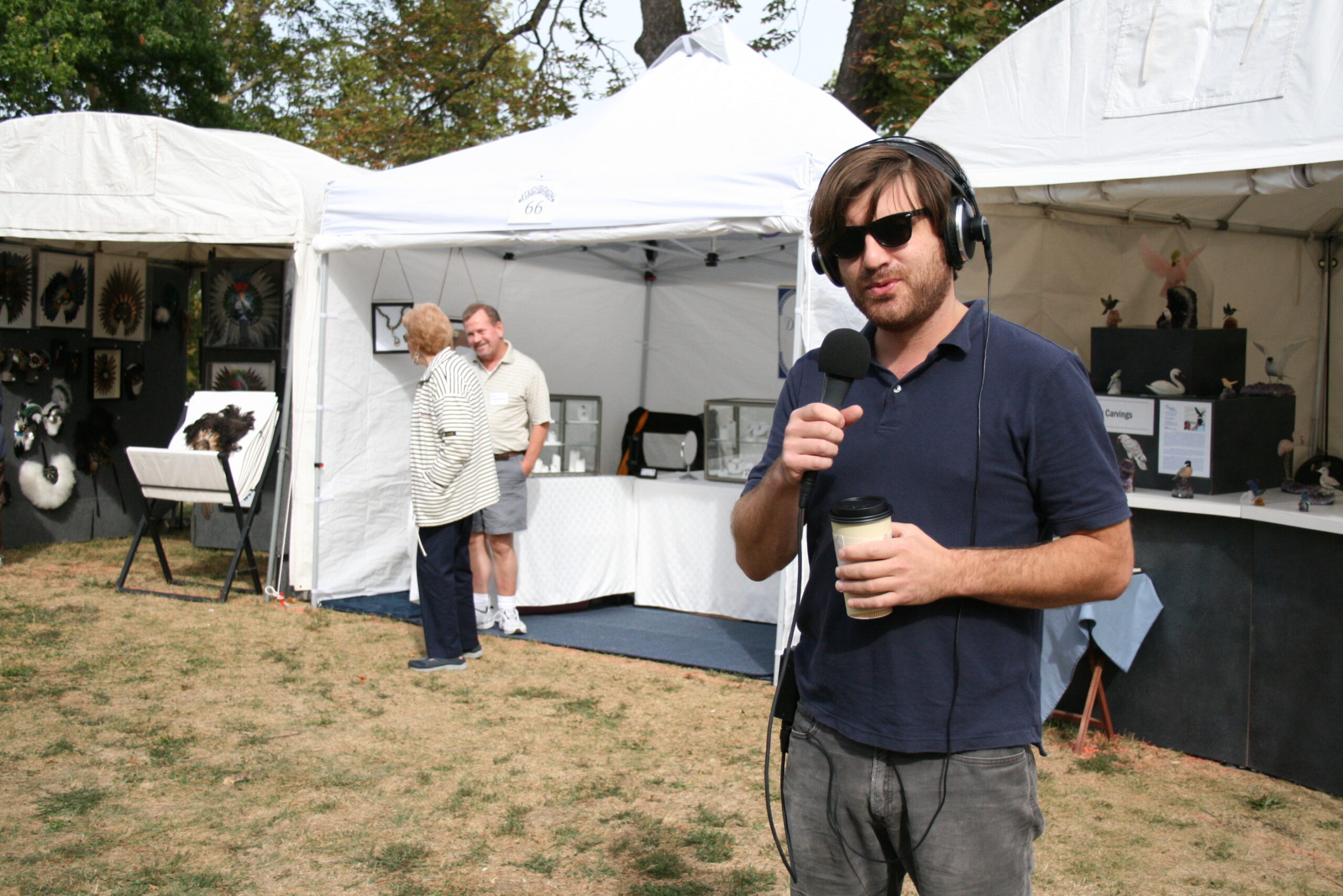 An adult wearing sunglasses and headphones and holding a microphone while standing in front of white tents