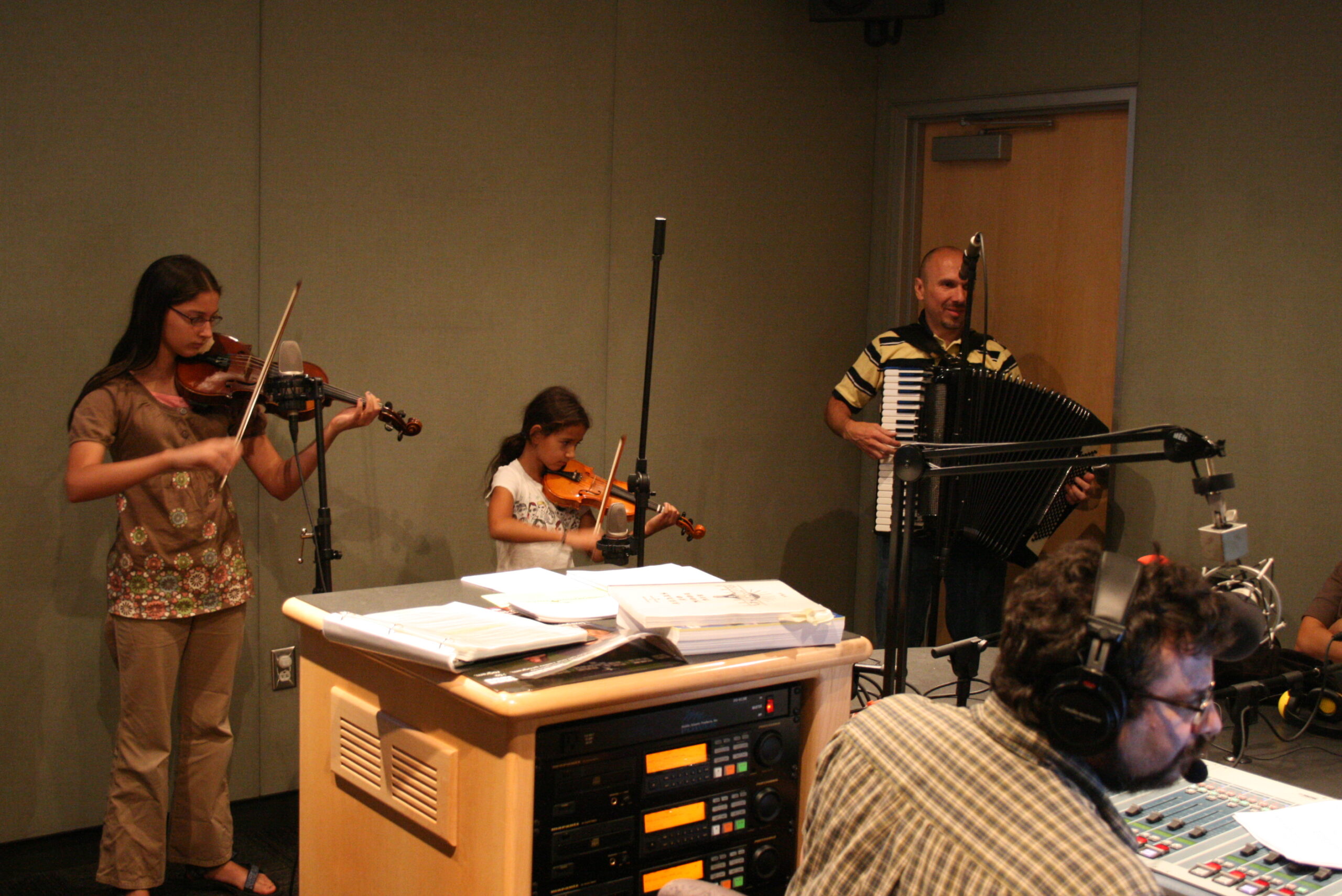 Two youth playing violins and an adult playing an accordion as an adult wearing headphones listens in a radio studio