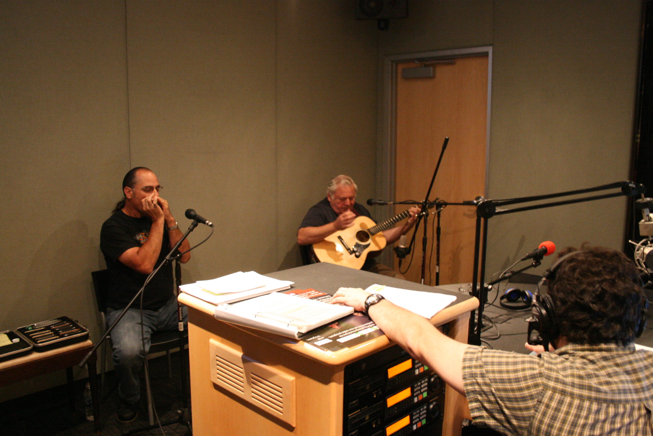 An adult playing a harmonica and an adult playing an acoustic guitar as an adult wearing headphones listens in a radio studio
