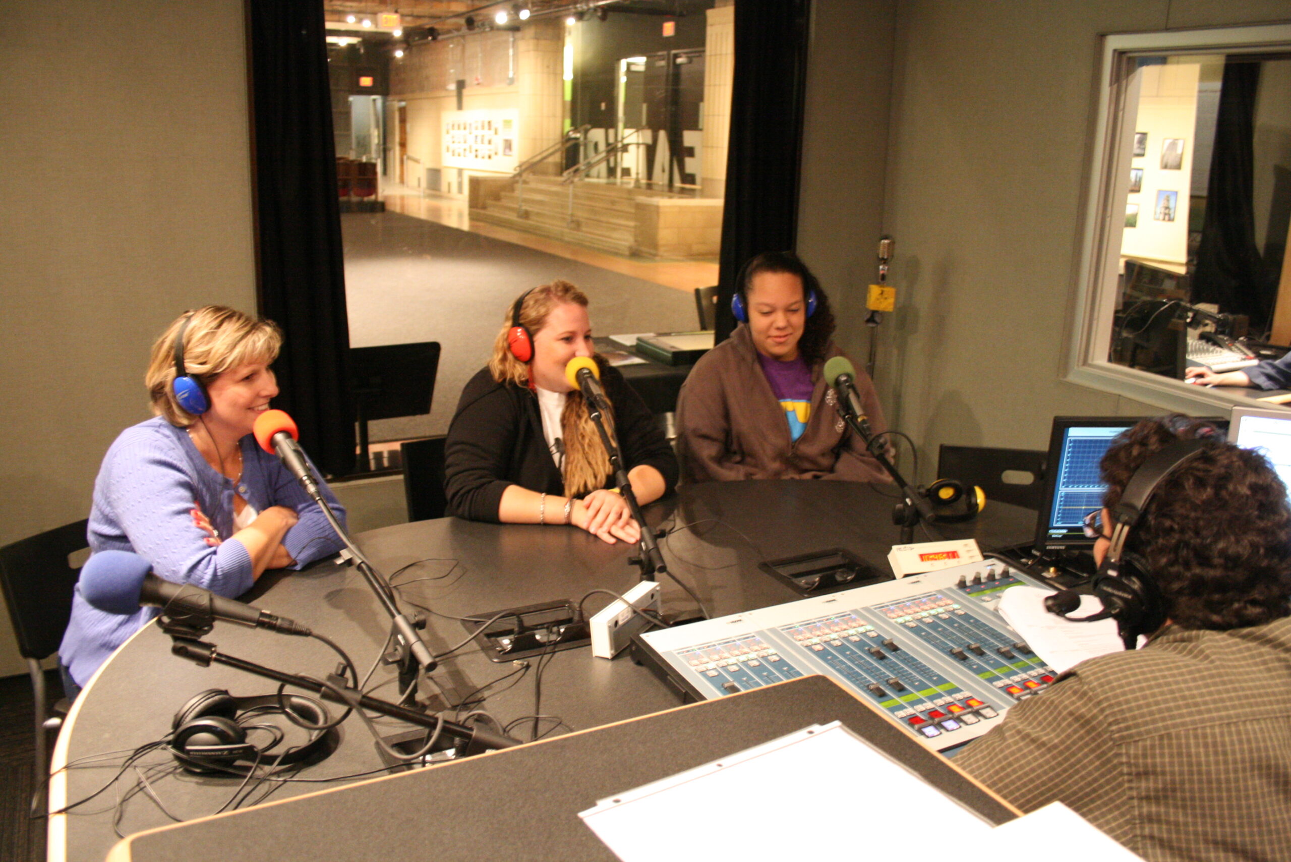 Two adults and a teen wearing headphones sitting behind microphones being interviewed by an adult wearing headphones sitting behind an audio mixer in a radio studio