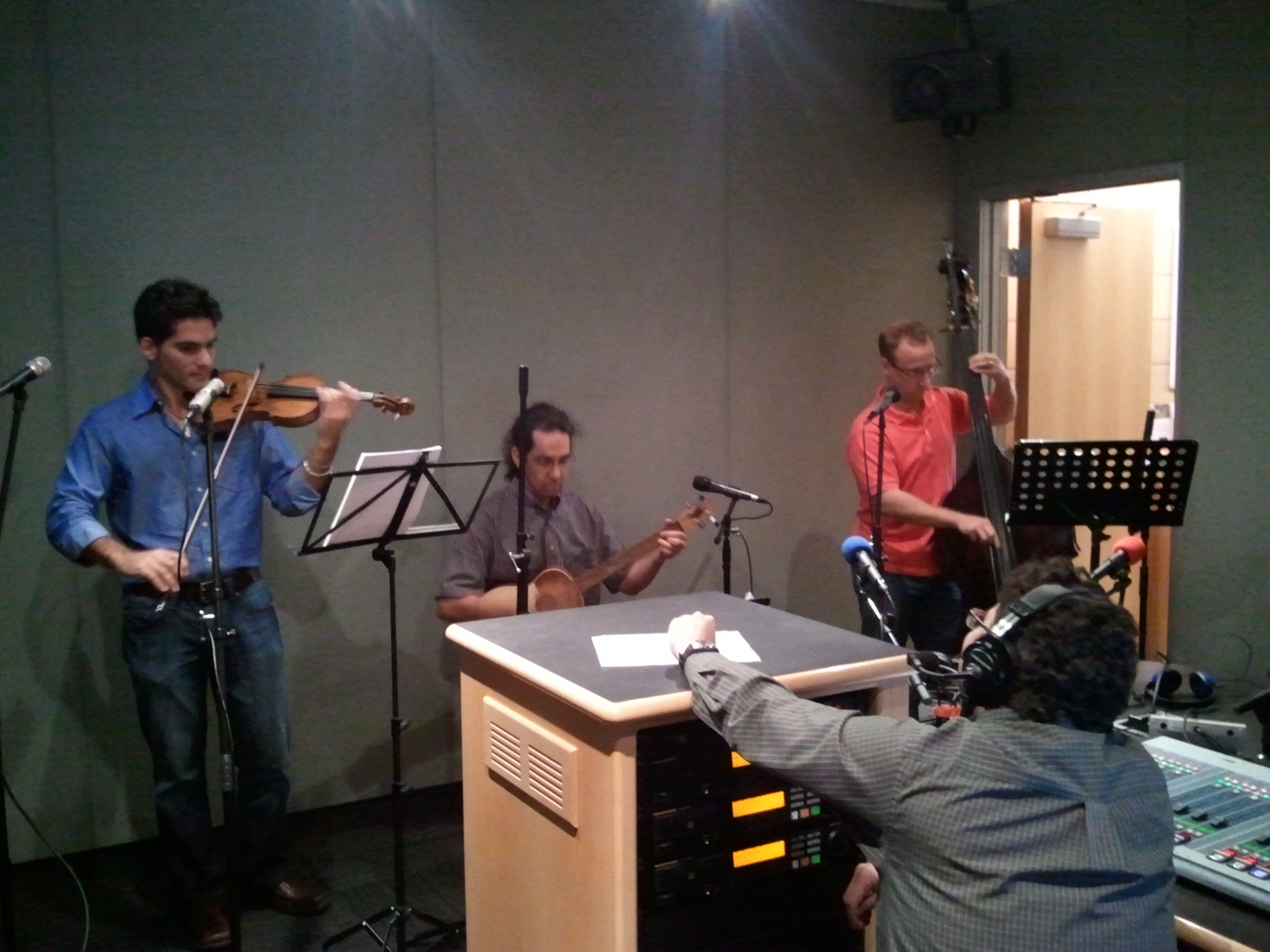 An adult playing a fiddle, an adult playing an acoustic guitar, and an adult playing an ethnic string instrument as an adult wearing headphones listens in a radio studio