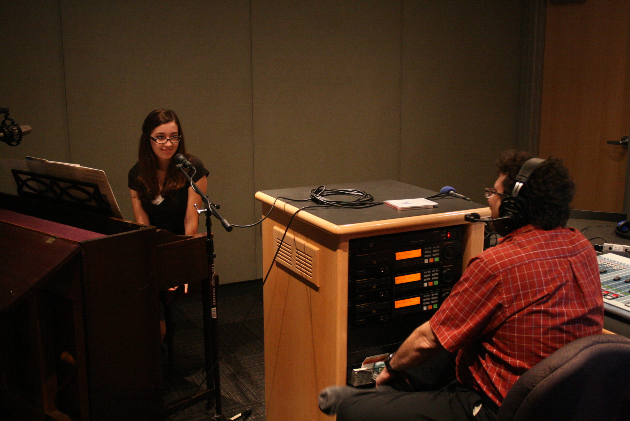 A teen sitting behind a piano being interviewed by an adult wearing headphones in a radio studio