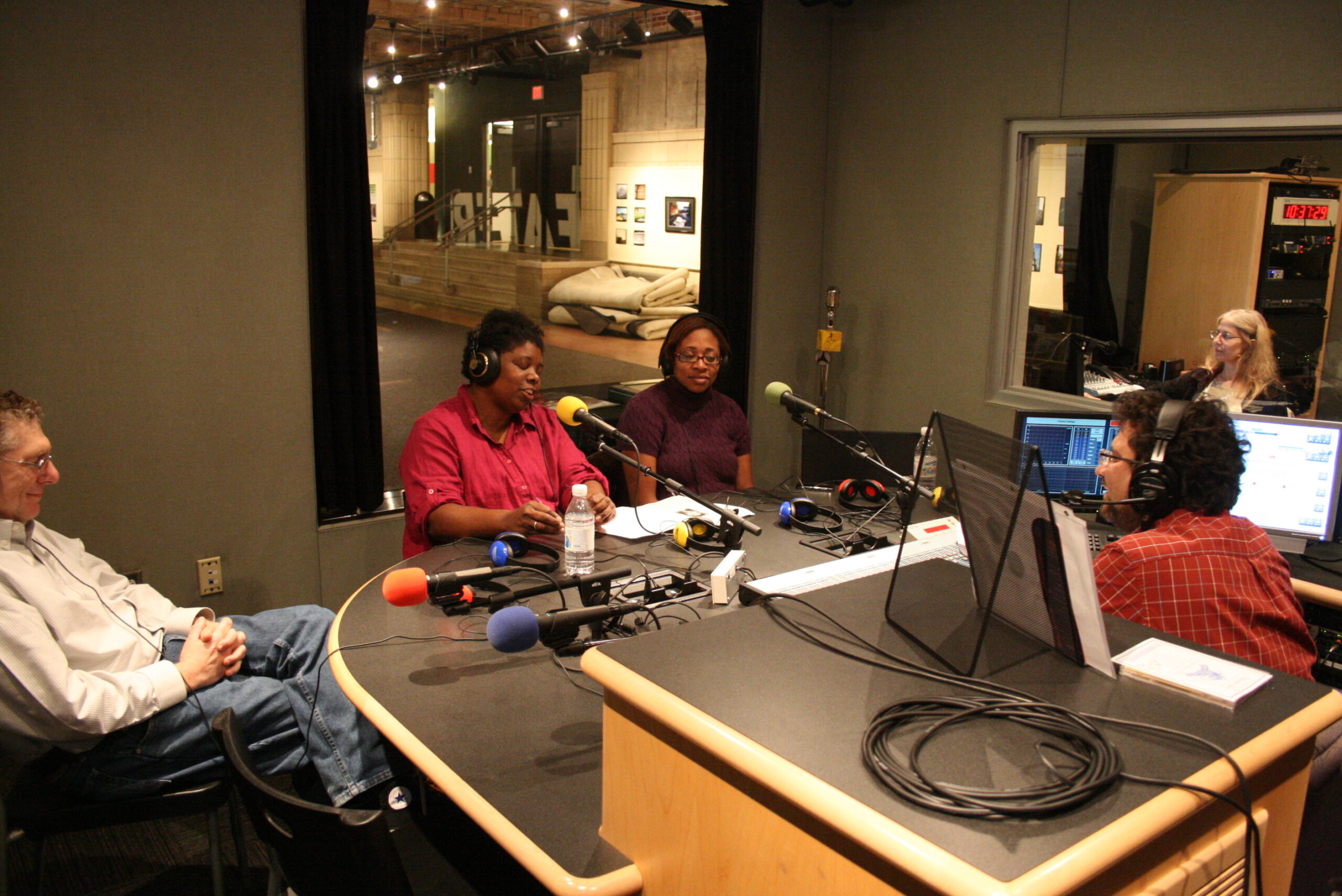Three adults wearing headphones speaking into microphones being interviewed by an adult wearing headphones sitting behind a microphone in a radio studio