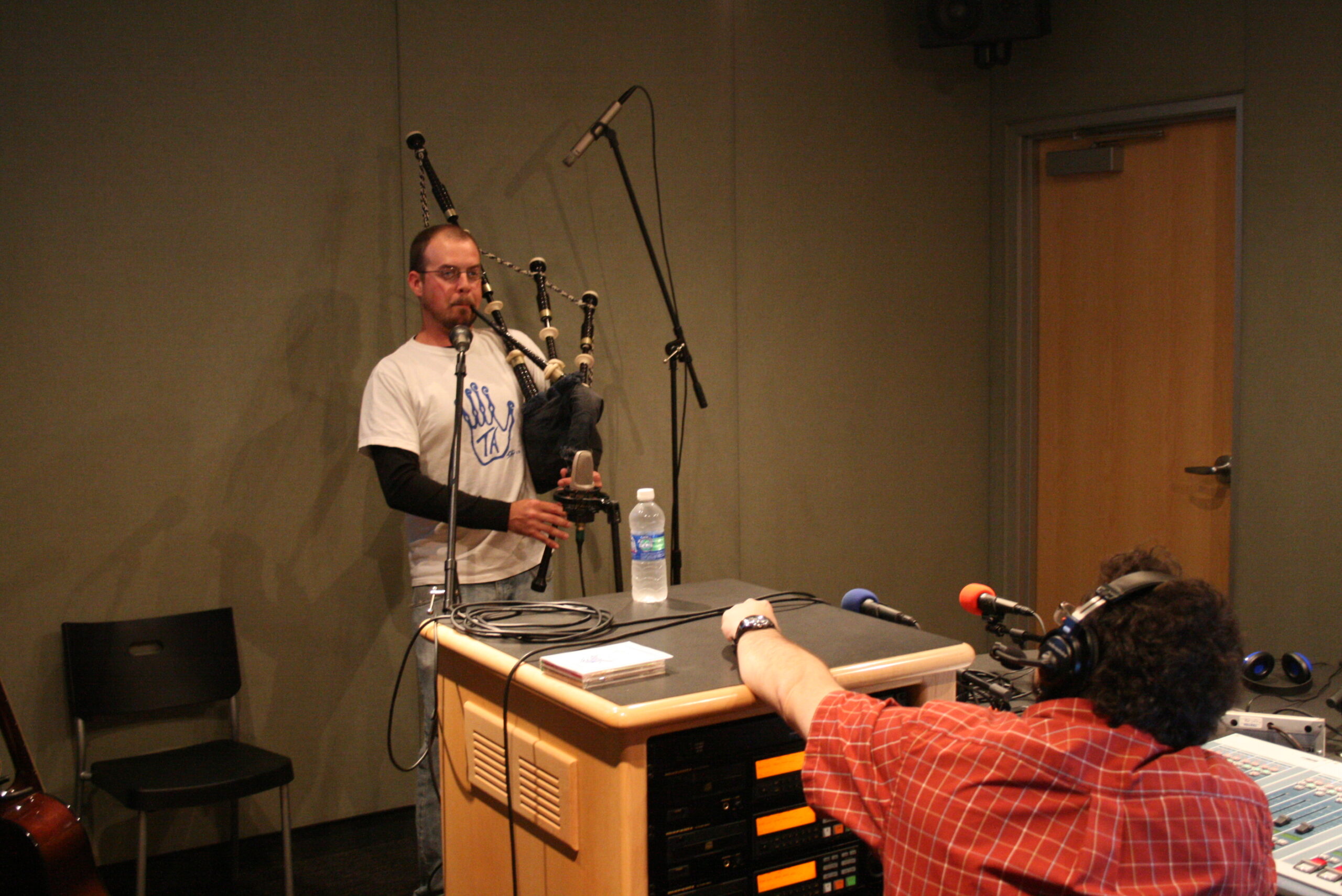 An adult playing a set of bagpipes as an adult wearing headphones listens in a radio studio