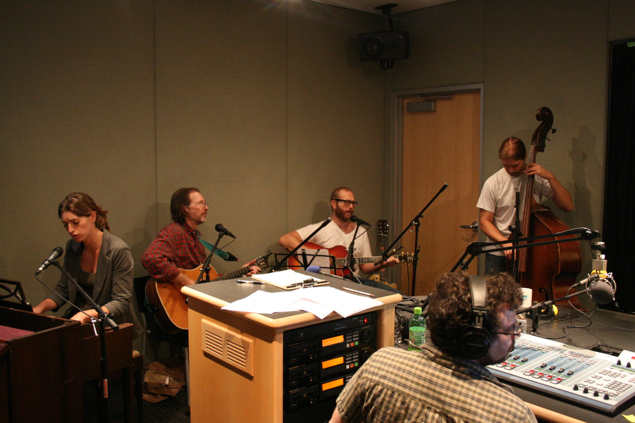 An adult playing a piano, two adults playing acoustic guitars, and an adult playing an upright bass as an adult wearing headphones listens in a radio studio