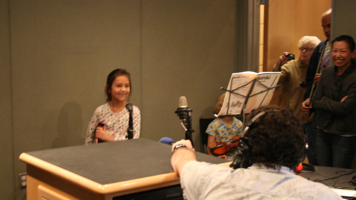A youth holding a violin and speaking into a microphone as an adult wearing headphones listens in a radio studio