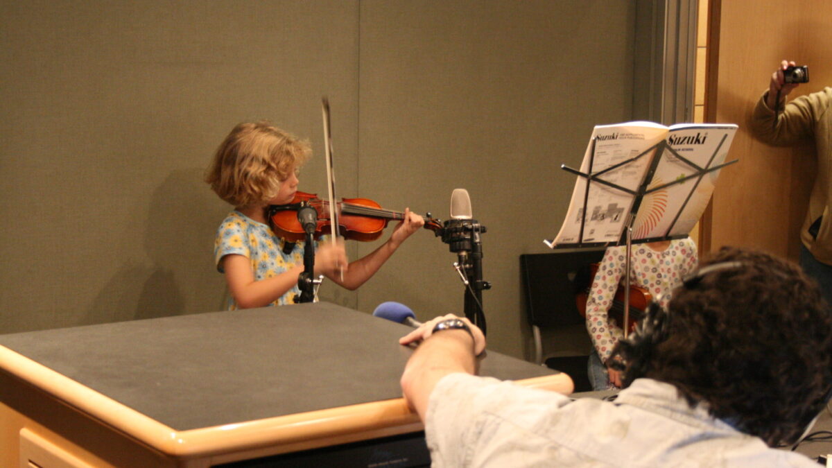 A youth playing a violin as an adult wearing headphones listens in a radio studio