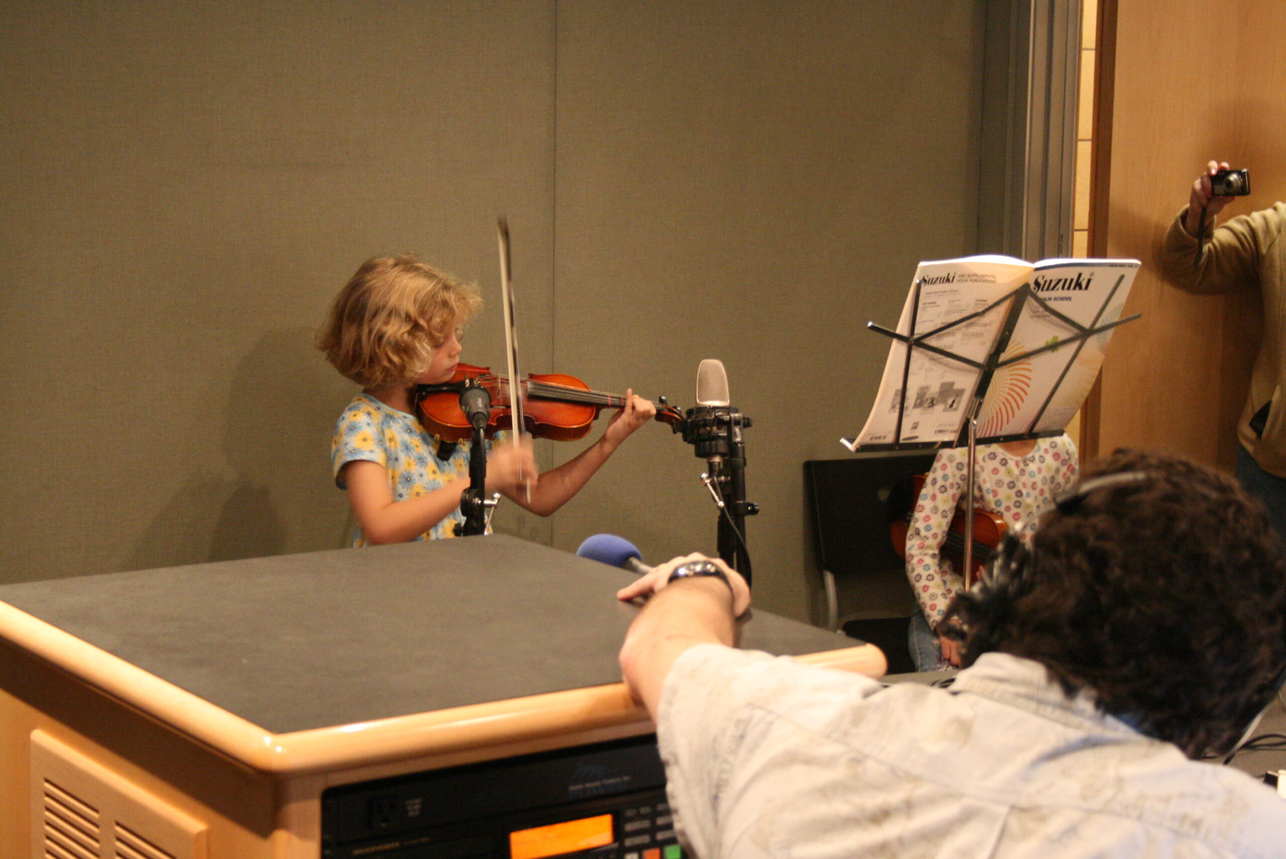 A youth playing a violin as an adult wearing headphones listens in a radio studio