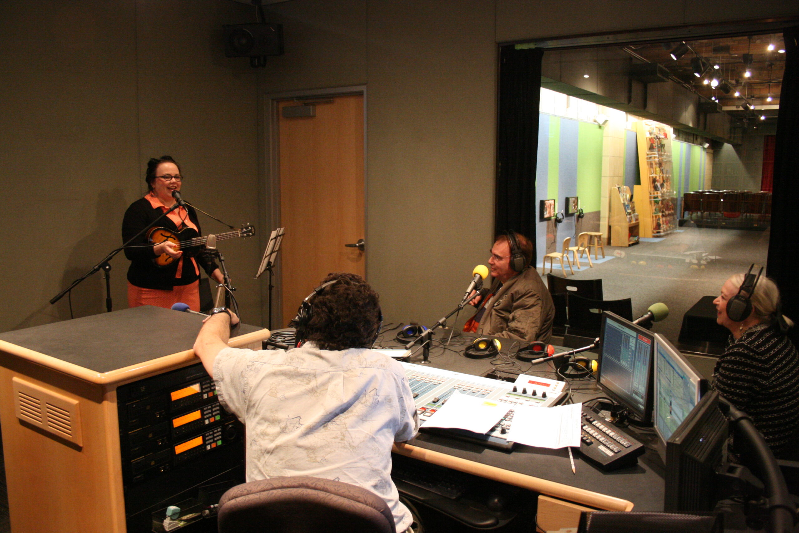 An adult holding a mandolin and speaking into a microphone as three adults wearing headphones listen in a radio studio.