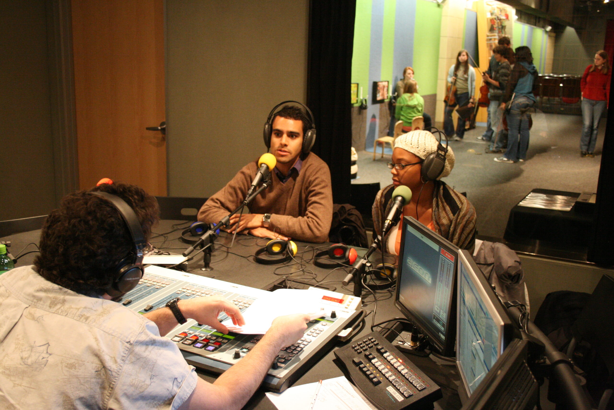Two teens wearing headphones speaking into microphones being interviewed by an adult wearing headphones sitting behind an audio mixer in a radio studio