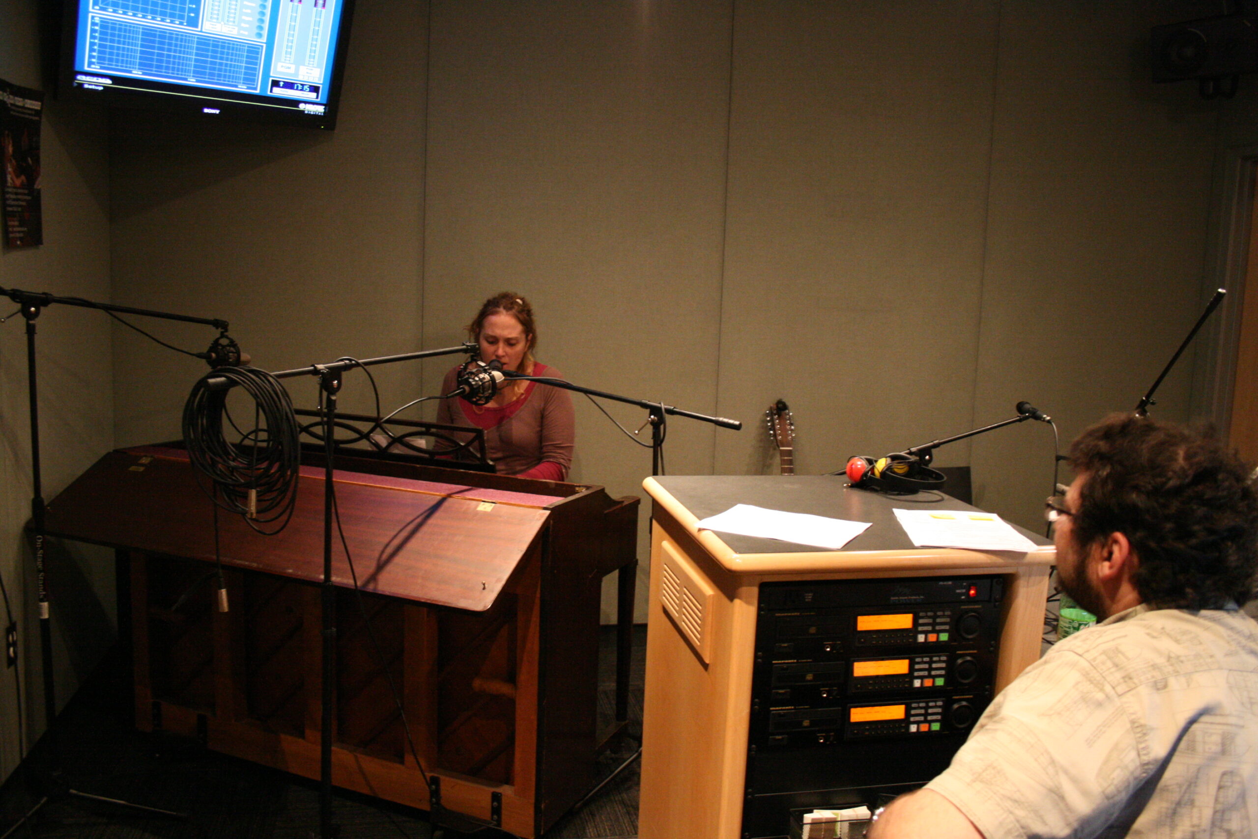 An adult playing a piano keyboard as an adult listens in a radio studio