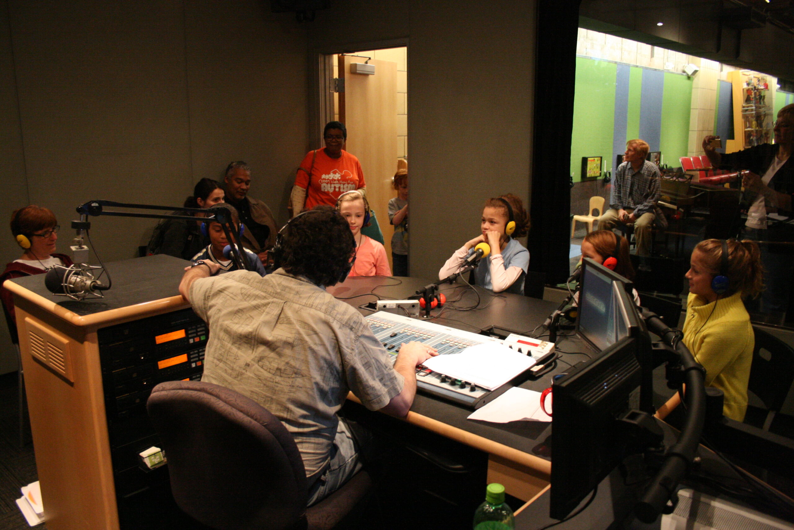 Six youth wearing headphones speaking into microphones being interviewed by an adult wearing headphones sitting behind an audio mixer in a radio studio