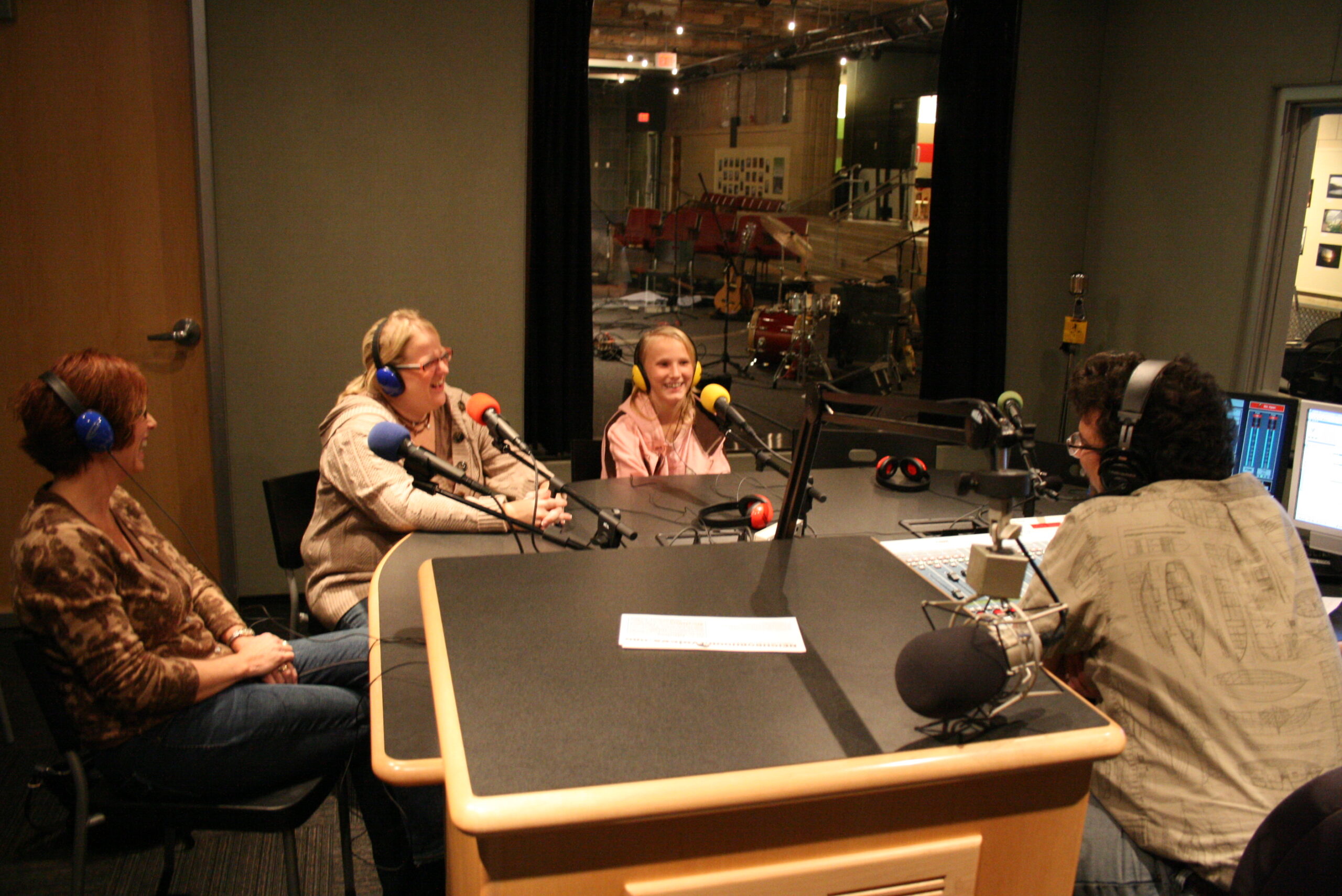 Two adults and a youth wearing headphones speaking into microphones being interviewed by an adult wearing headphones sitting behind an audio mixer and a microphone in a radio studio