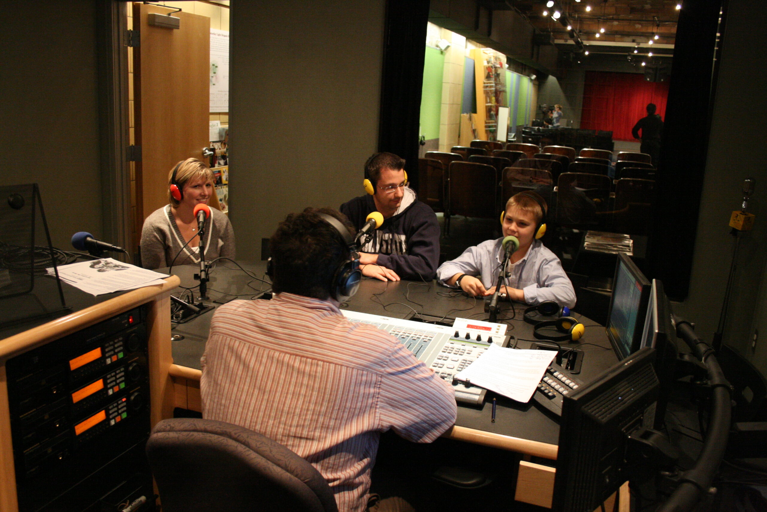 Two adults and a youth wearing headphones speaking into microphones being interviewed by an adult sitting behind an audio mixer in a radio studio