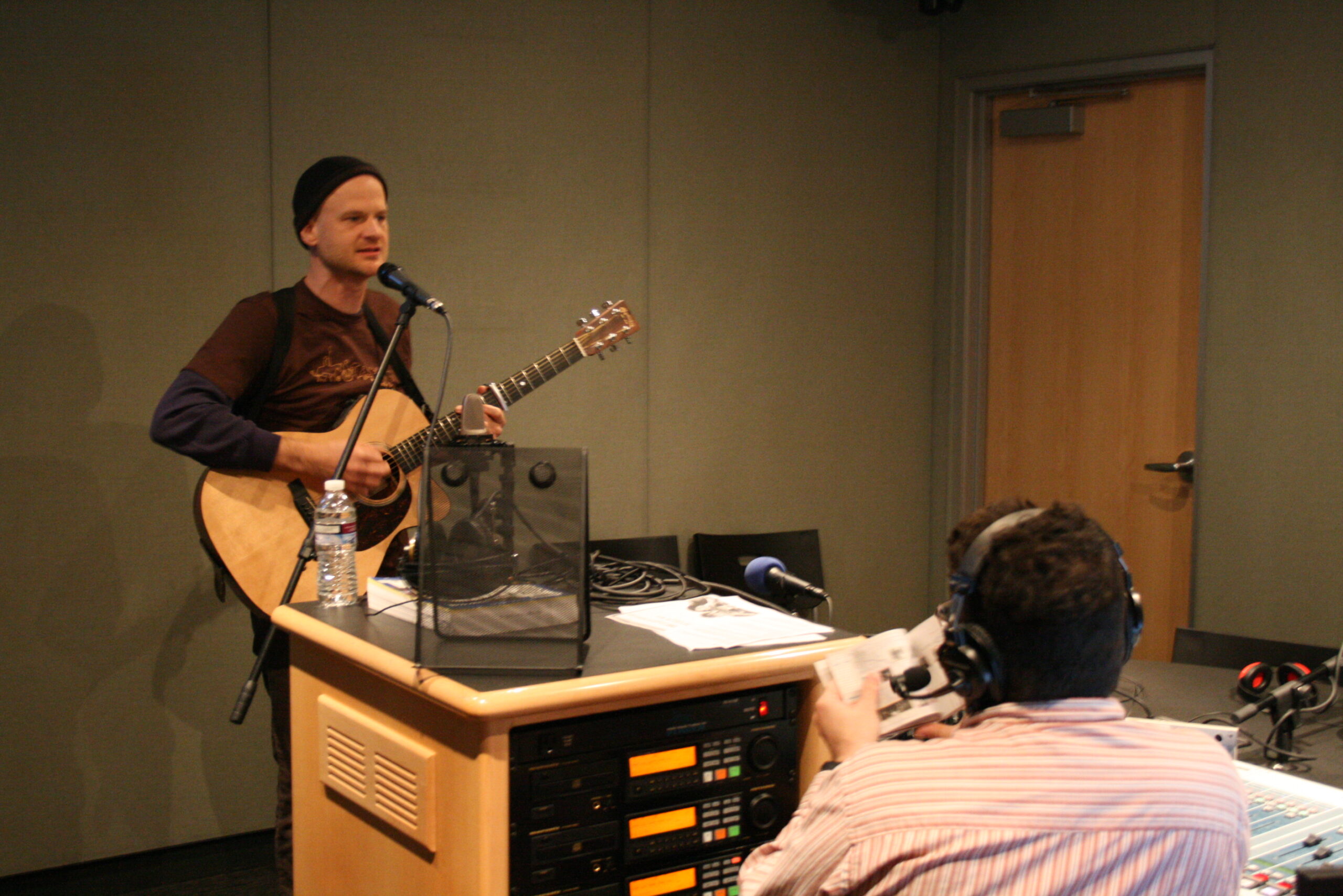 An Adult holding an acoustic guitar and speaking into a microphone being interviewed by an adult wearing headphones in a radio studio
