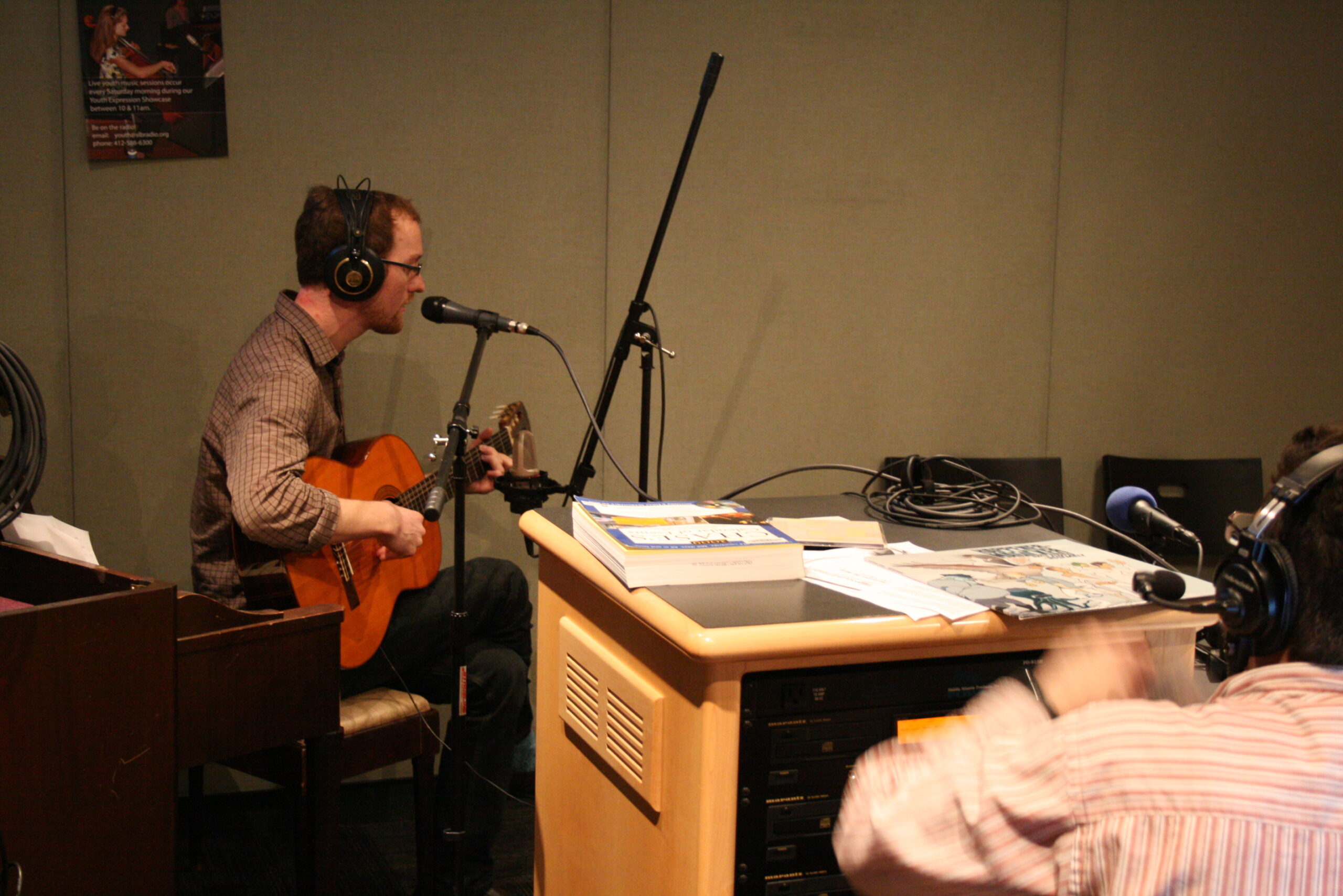 An adult wearing headphones playing an acoustic guitar and singing into a microphone as an adult wearing headphones listens in a radio studio