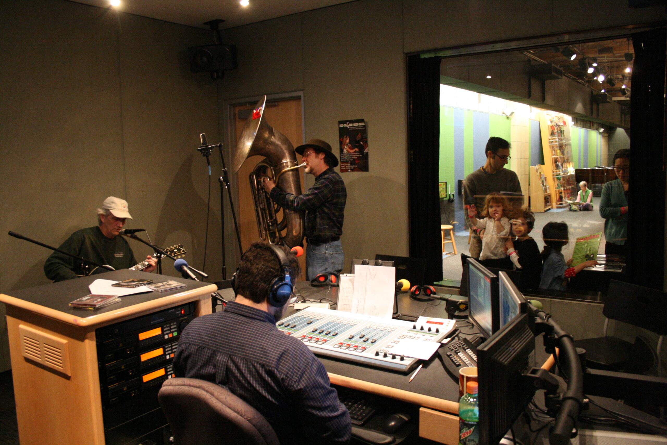 An adult playing an acoustic guitar and an adult playing a tuba as an adult sitting behind an audio mixer in a radio studio