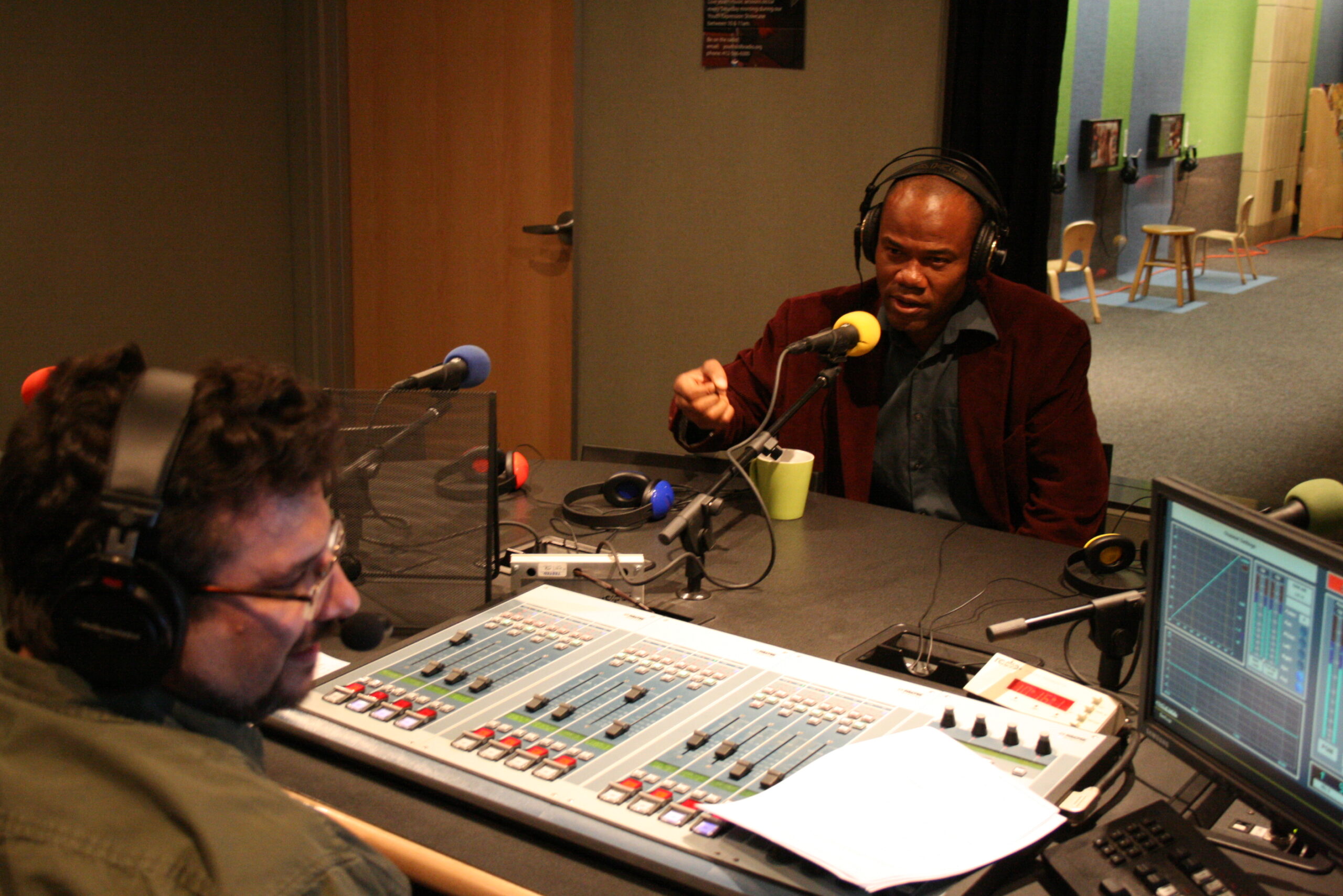 An adult wearing headphones speaking into a microphone being interviewed by an adult wearing headphones sitting behind an audio mixer in a radio studio