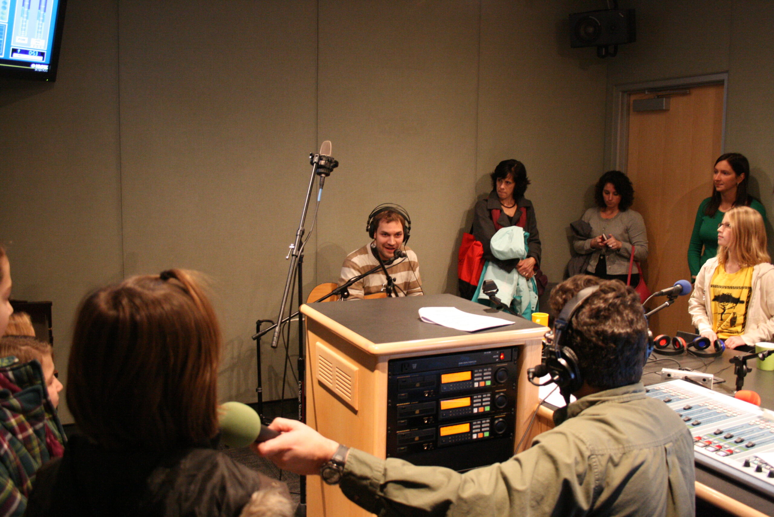 An adult playing an acoustic guitar and singing into a microphone and six adults singing in a choir as an adult wearing headphones listens in a radio studio