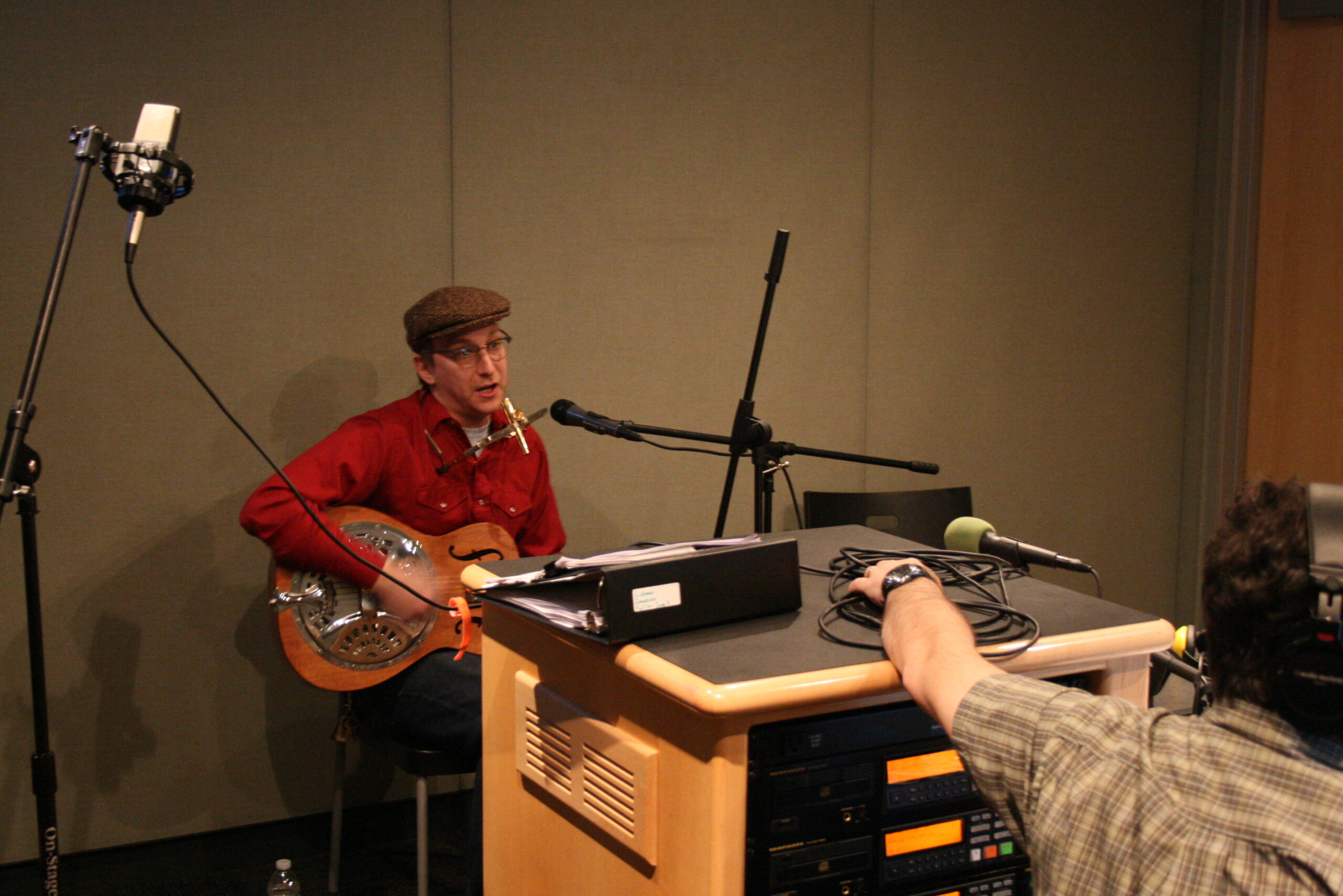 An adult playing a dobro and singing into a microphone as an adult wearing headphones listens in a radio studio
